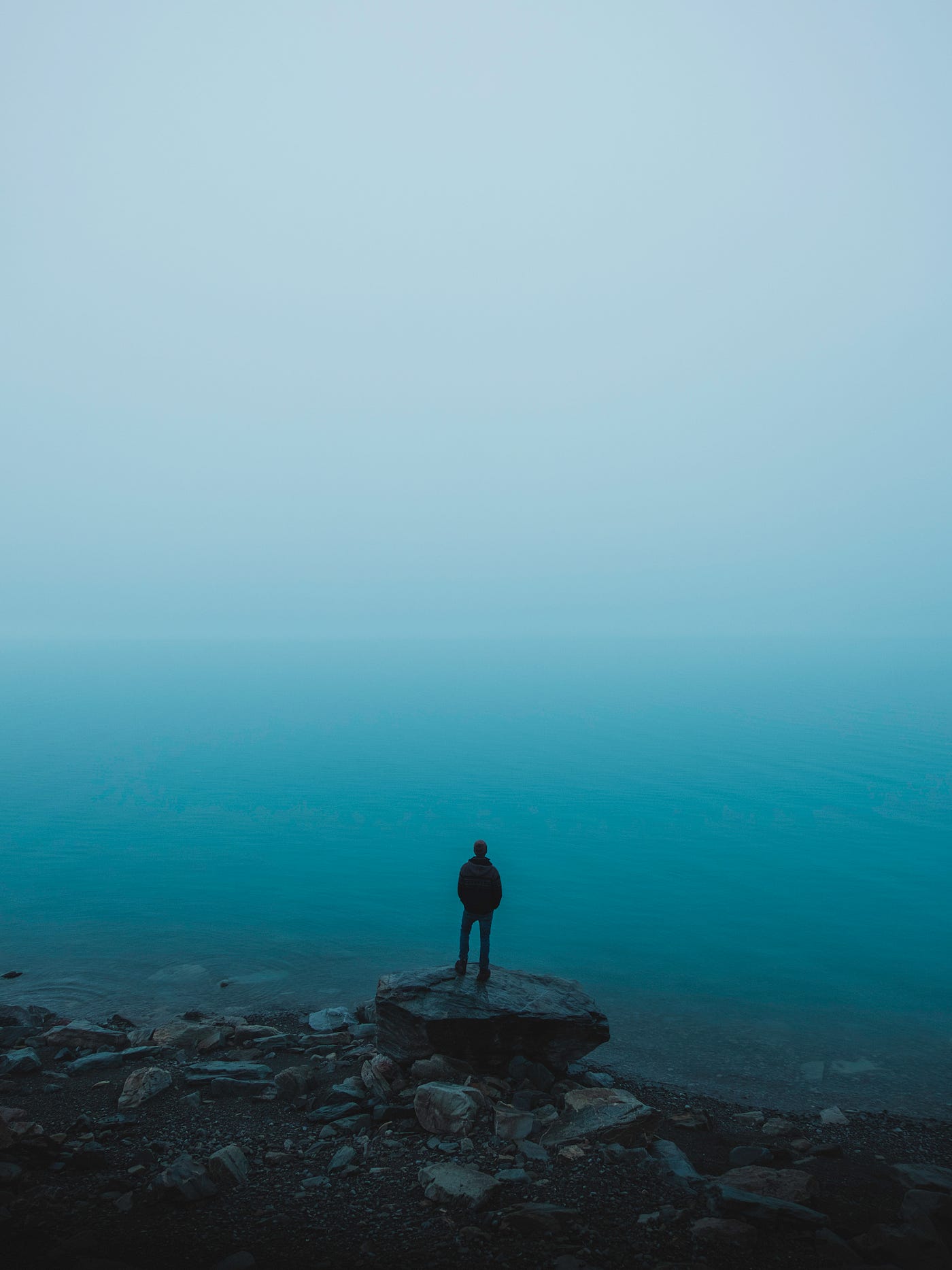 A man stands on a rock, looking at the ocean in the distance. The scene is a hazy blue.