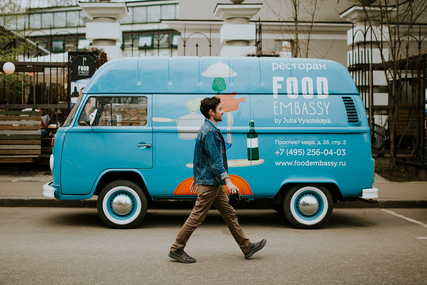 A young man, clad in a jeans jacket and brown pants, walks to the right. Behind him is a light blue food truck with writing on it advertising “Food Embassy.”