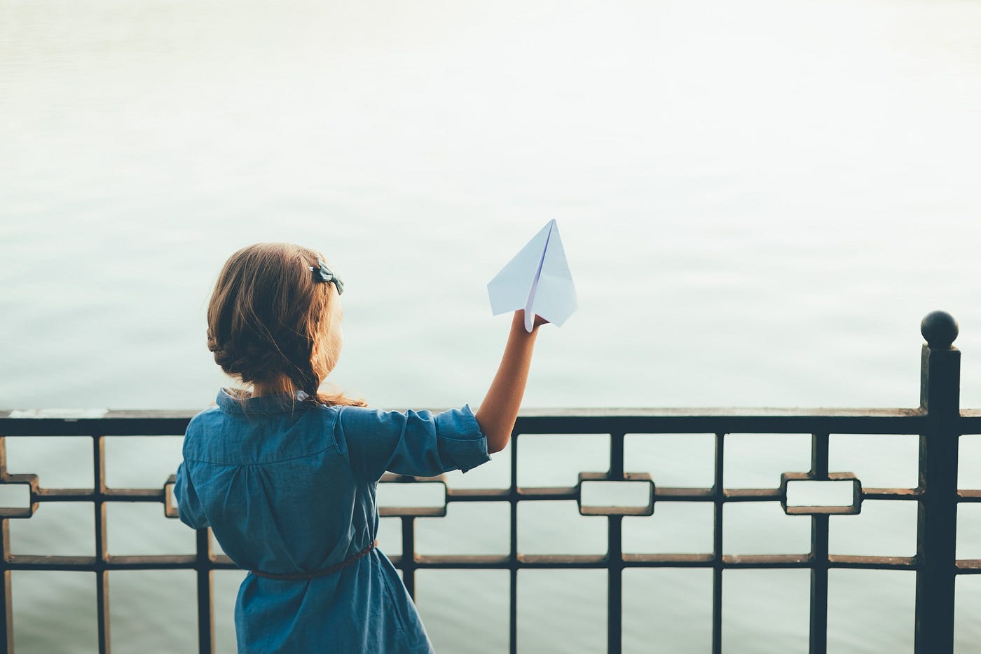 Girl in blue dress launching toy paper airplane looking to lake