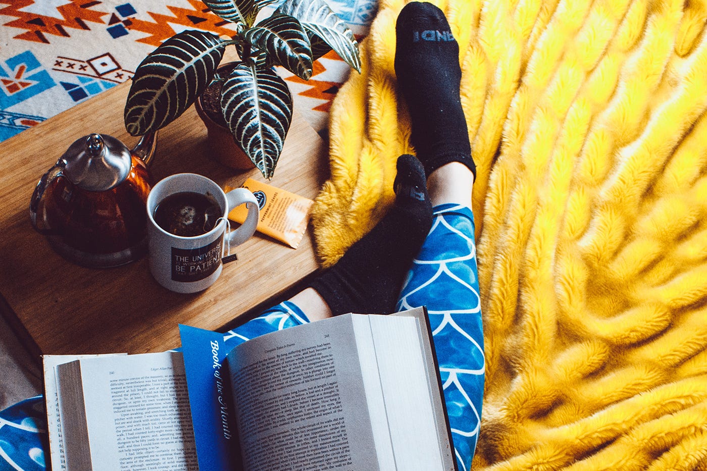 A person’s legs and feet are visible as they lay in bed reading a book, with a tray with a teapot, mug, and a plant alongside