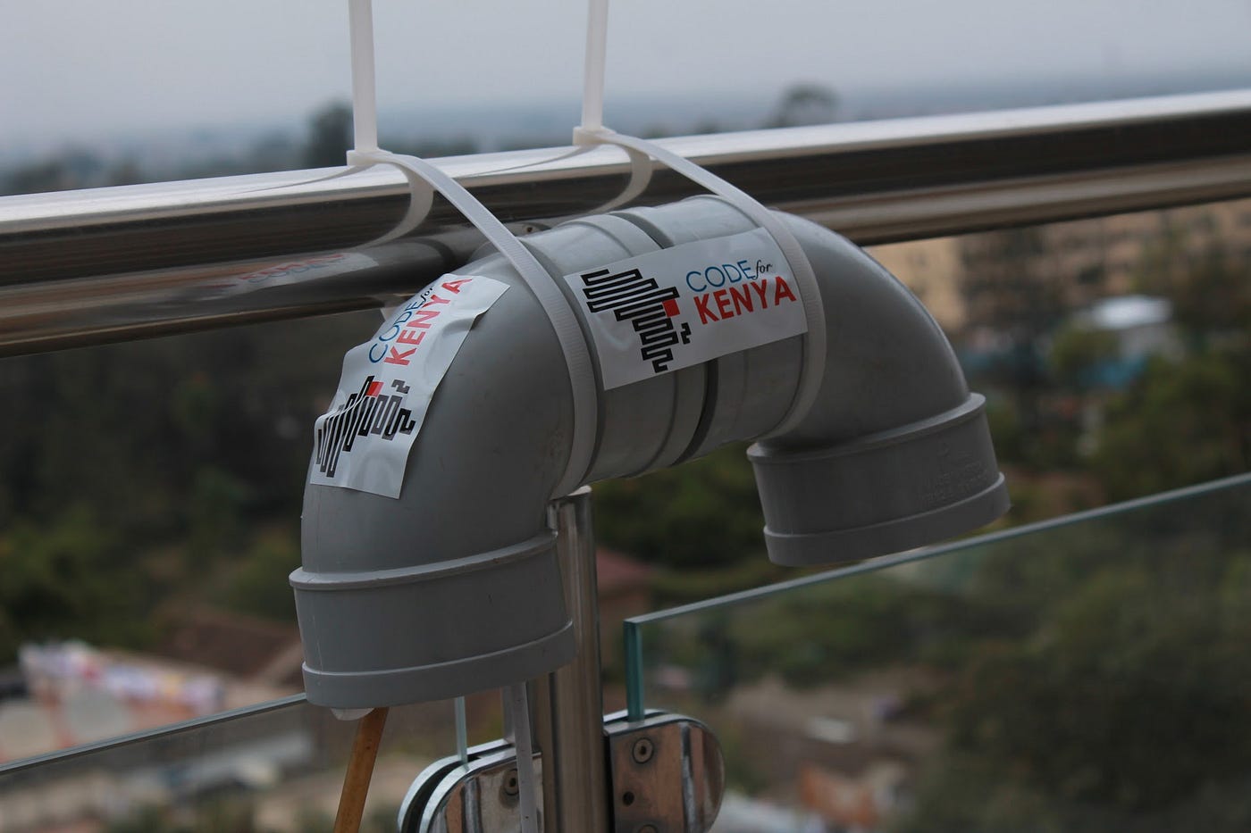An air quality sensor outdoors on the railing of a building. The Code for Kenya logo is visible on the sensor.