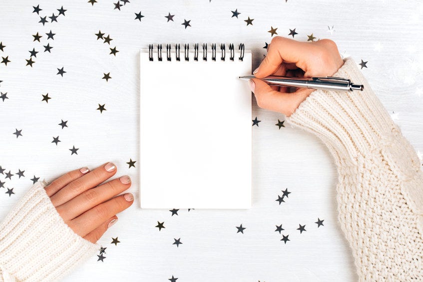 A woman’s hands against a white background with gold starts holds a pen over a blank notebook page.