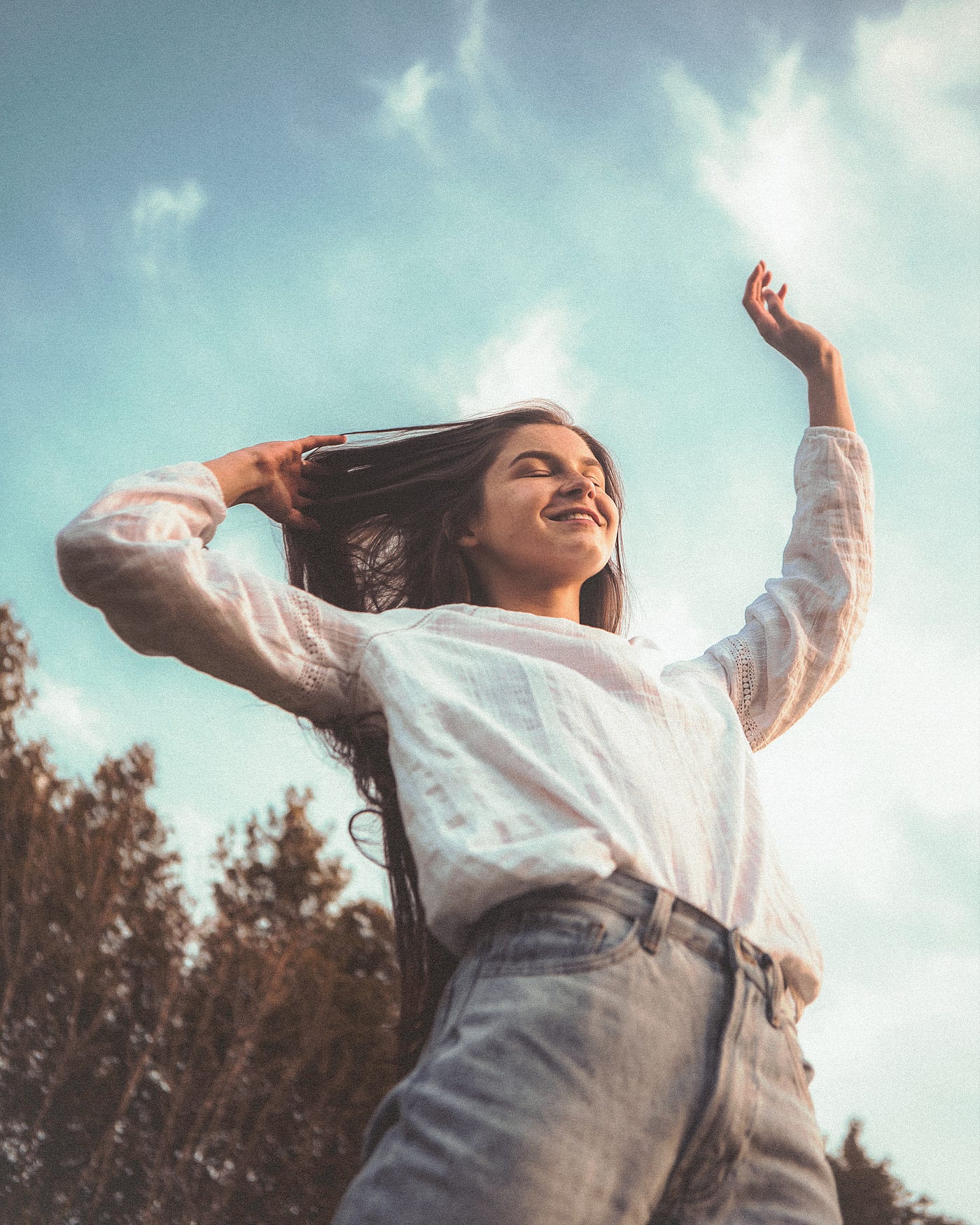 Young woman, seen from the waist up, extends her left arm overhead in joy. Her bent right arm extends upwards too. Blue sky in background with a few trees in the distance.