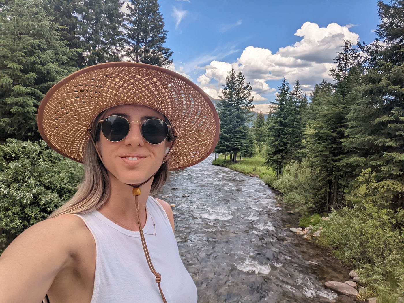 A woman in a wide brimmed hat taking a selfie in front of a river. There are trees on either side of the river. She is wearing sunglasses and a white tank top.