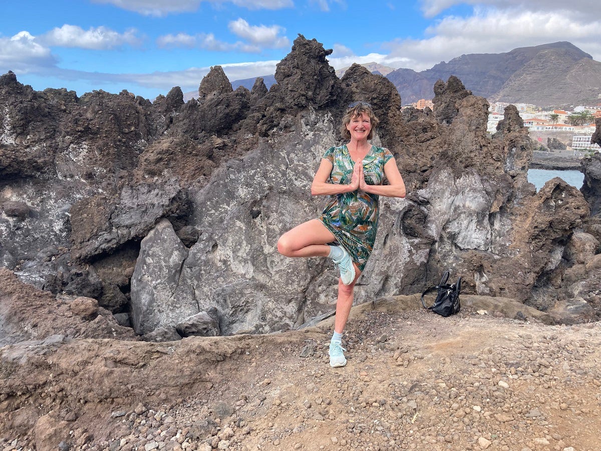 I’m standing on one leg in this picture (I’m a woman in my fifties). There are volcanic rocks behind — this is Tenerife — and cliffs in the distance, and a glimpse of blue sea.