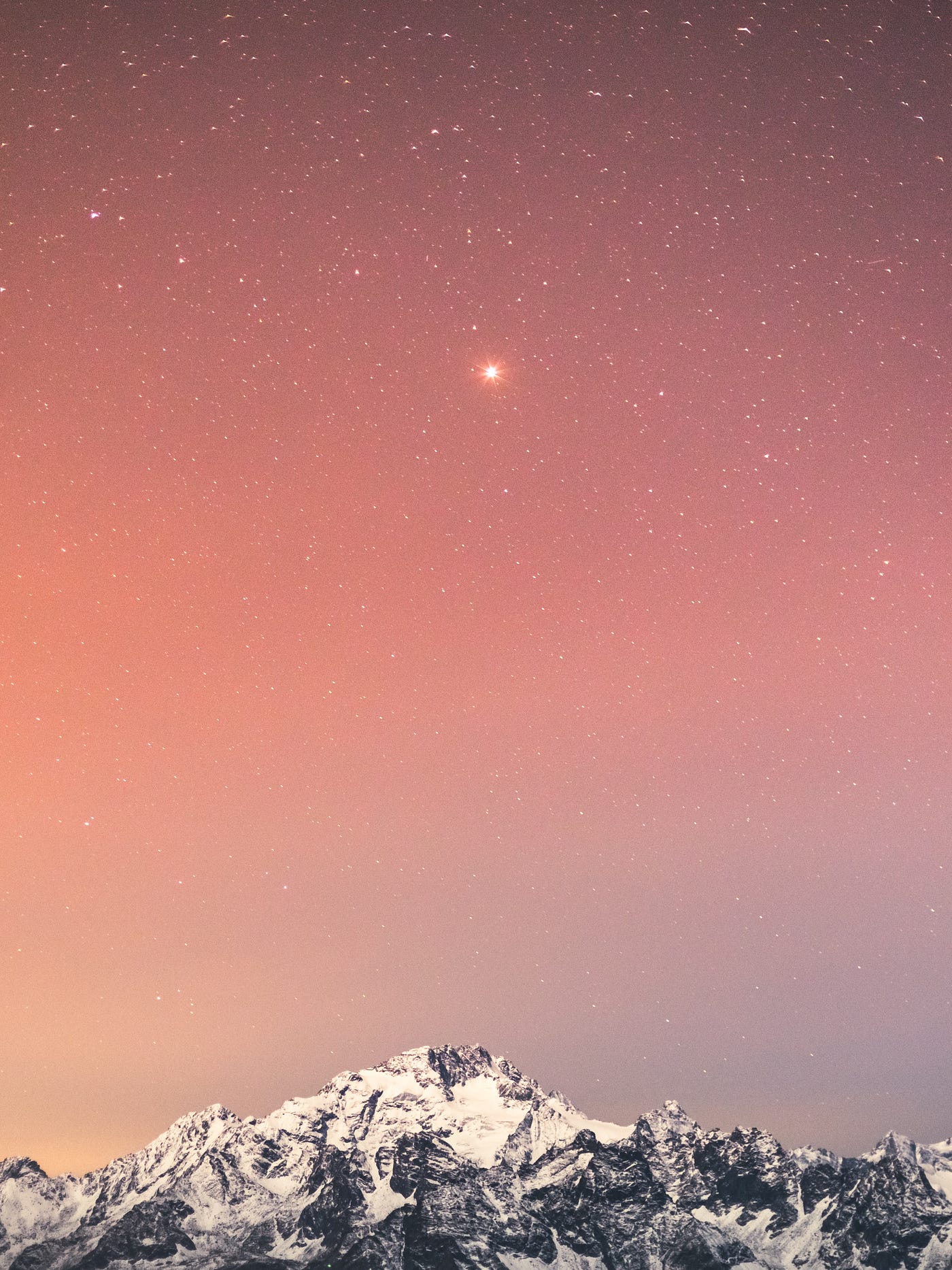 One bright star amidst many on a pink sunset sky above a craggy mountaintop