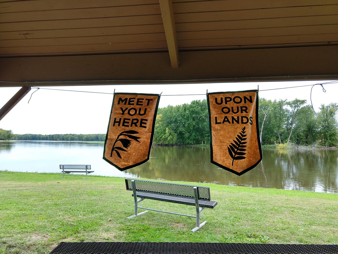Two banners reading “Meet you here” and “Upon our lands” in picnic shelter near river.