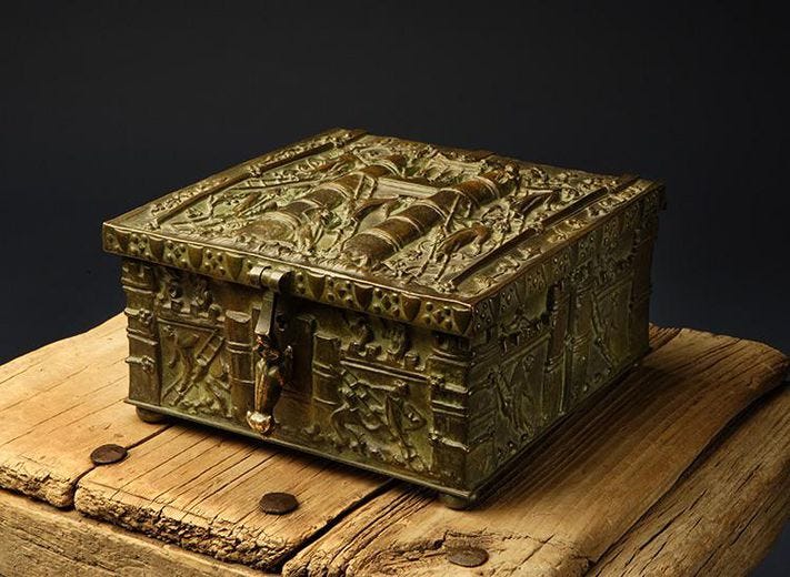 A brass-colored metal box with ornate inscription sits closed on a wooden platform.