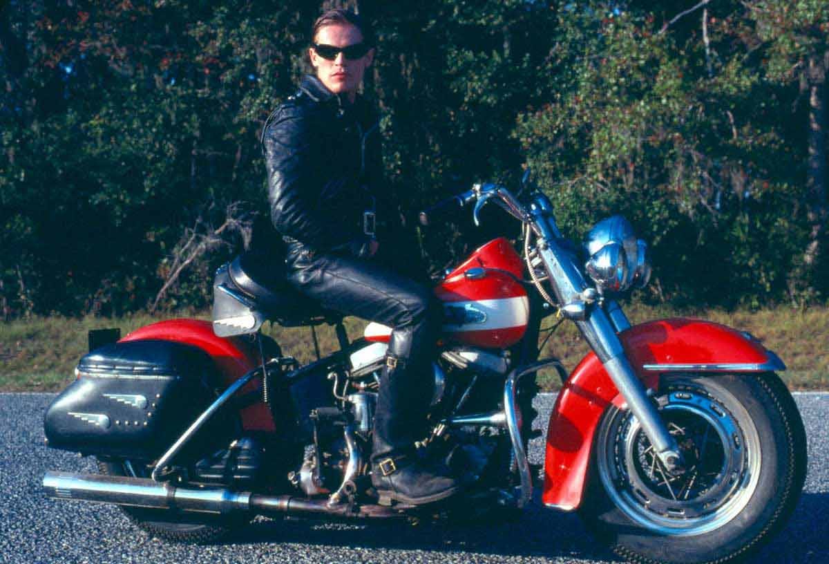 Willem Dafoe sits on an vintage motorcycle.