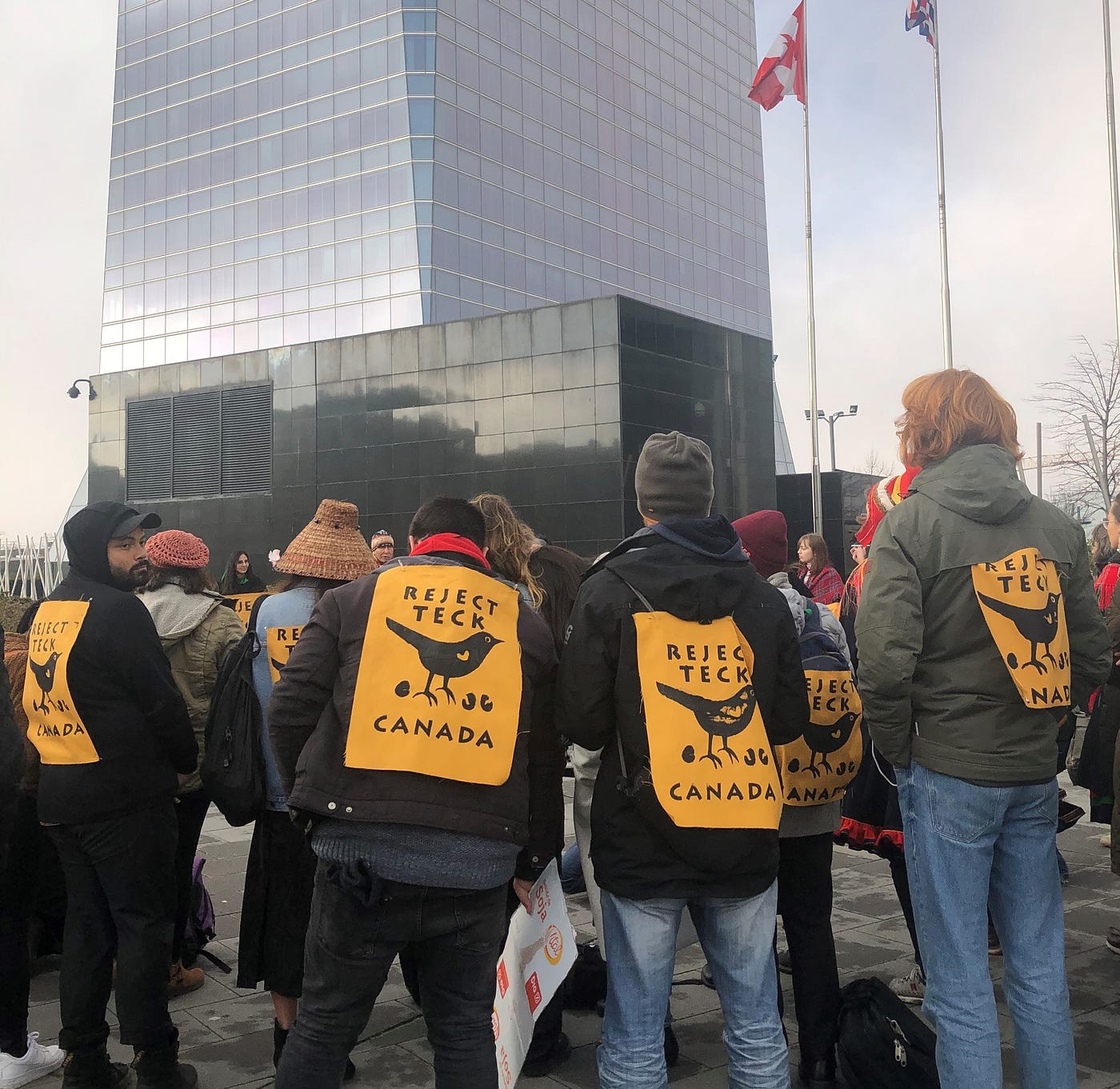 Four activists standing outside of the Canadian embassy building in Madrid, Spain, with signs on their backs “Reject Teck”