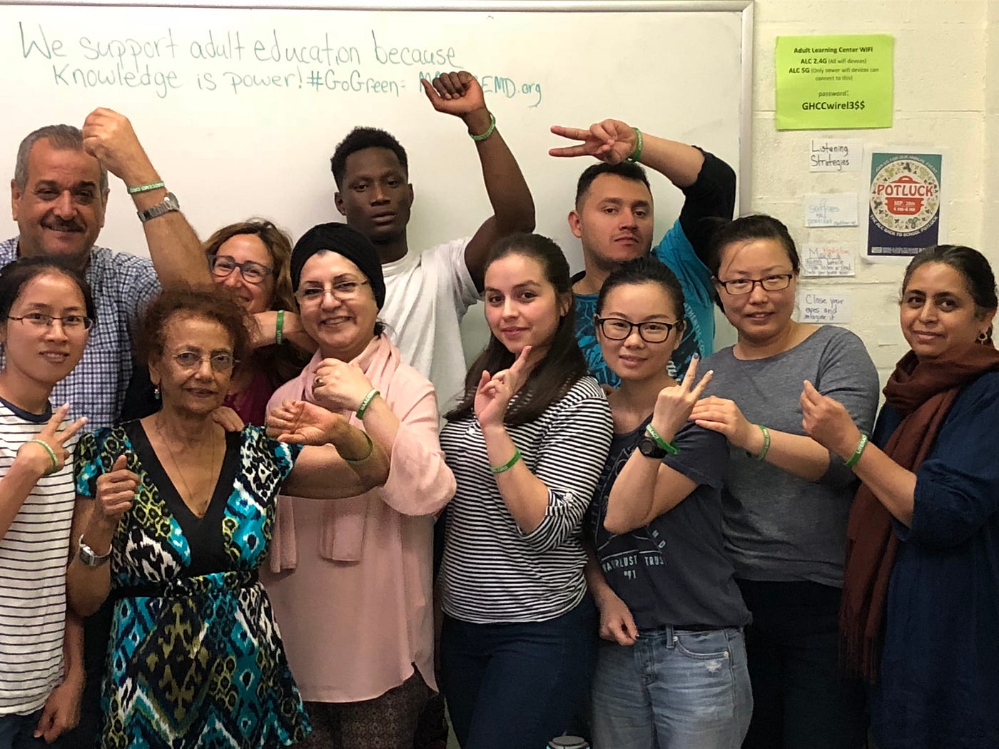 A smiling group of 13 students pose in a classroom with fists and peace hand signs raised.