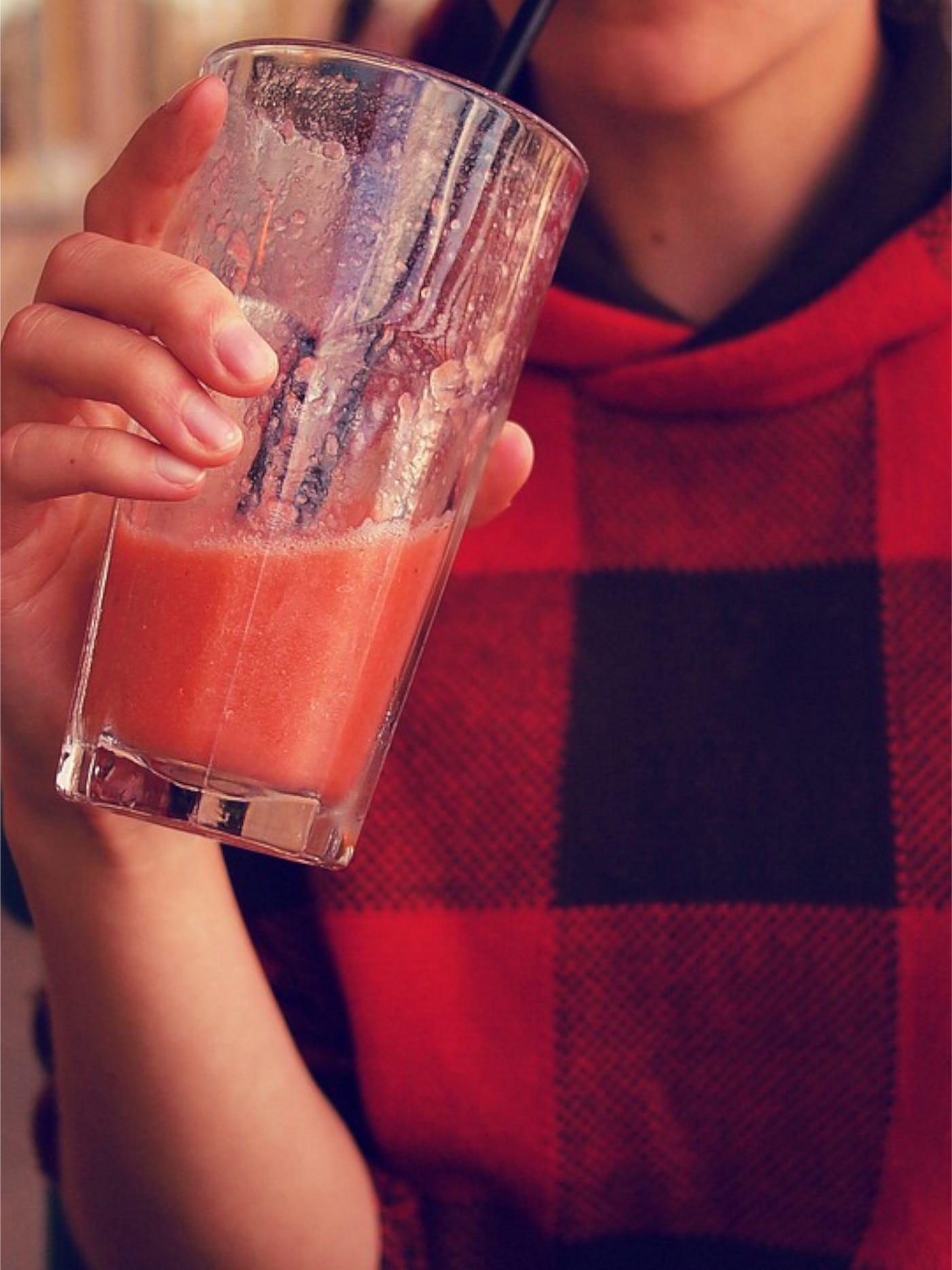 Color photo of a woman wearing a red buffalo plaid top while drinking a red beverage.