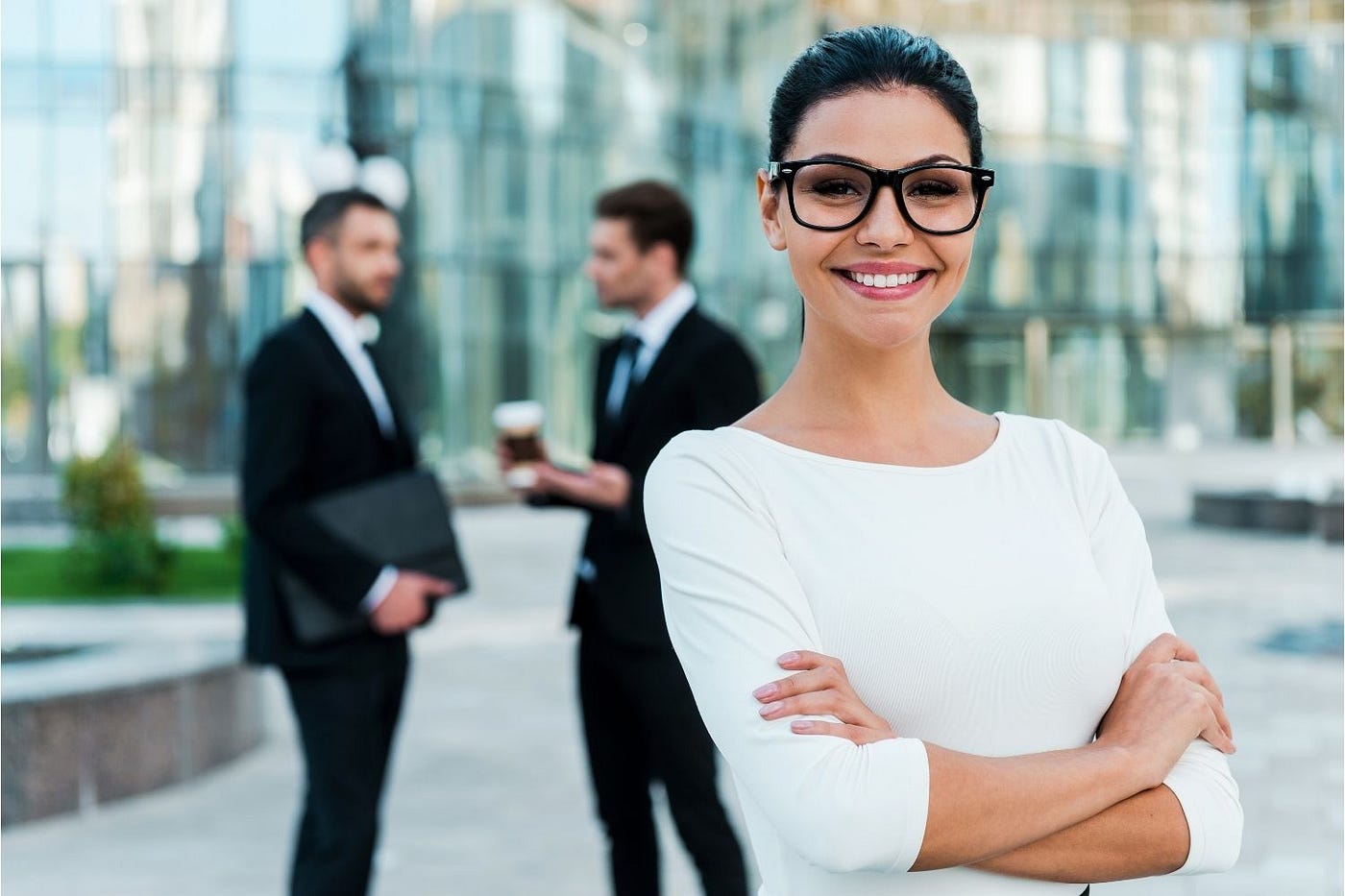 Businesswoman standing in the foreground with arms crossed and smiling confidently with two businessmen in the background.