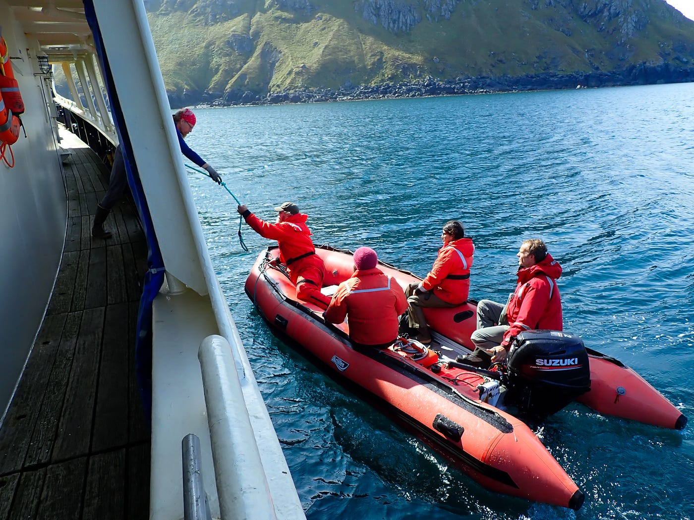 An orange rubber skiff with 4 people in orange safety suits pulls up to a larger boat in the ocean.