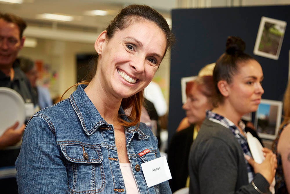 A smiling mature student attending an event on campus. There are several mature students looking at displays of work and photography.