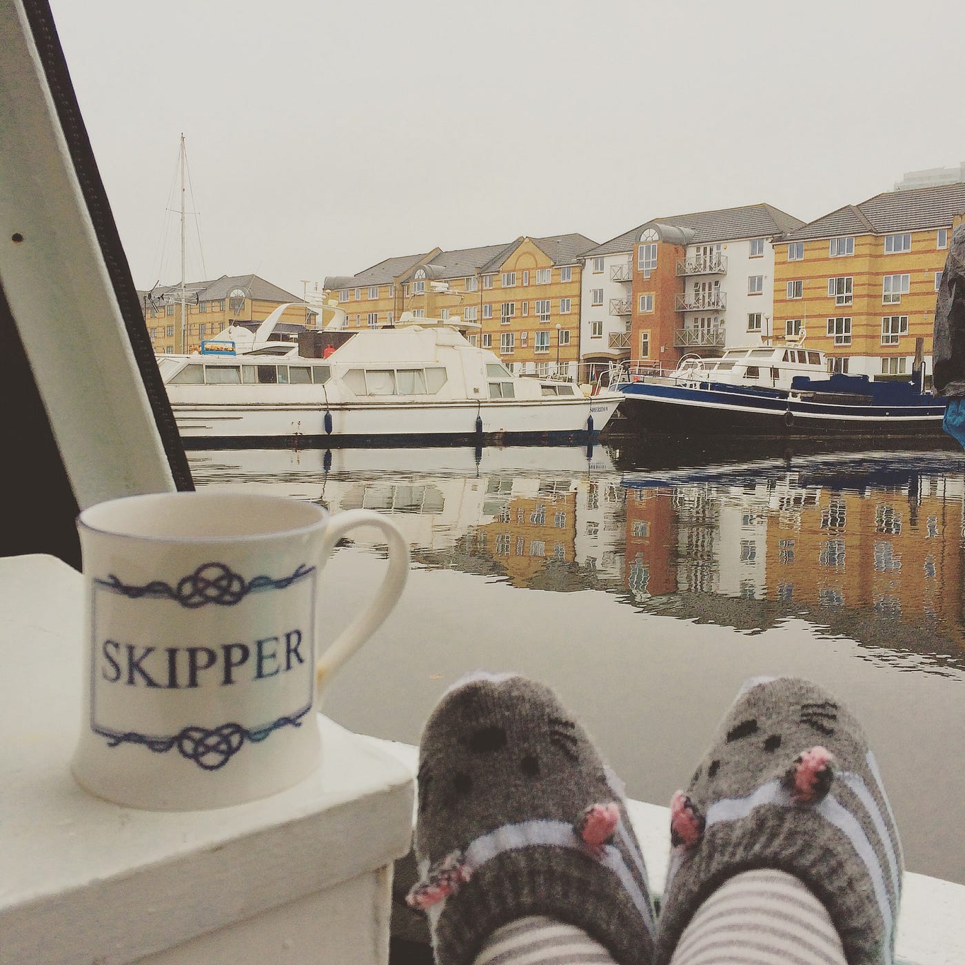 Photo showing the view of a boat yard on a rainy day, with warm grey slippers propped up on a rail and a mug that says ‘skipper’ on the outside