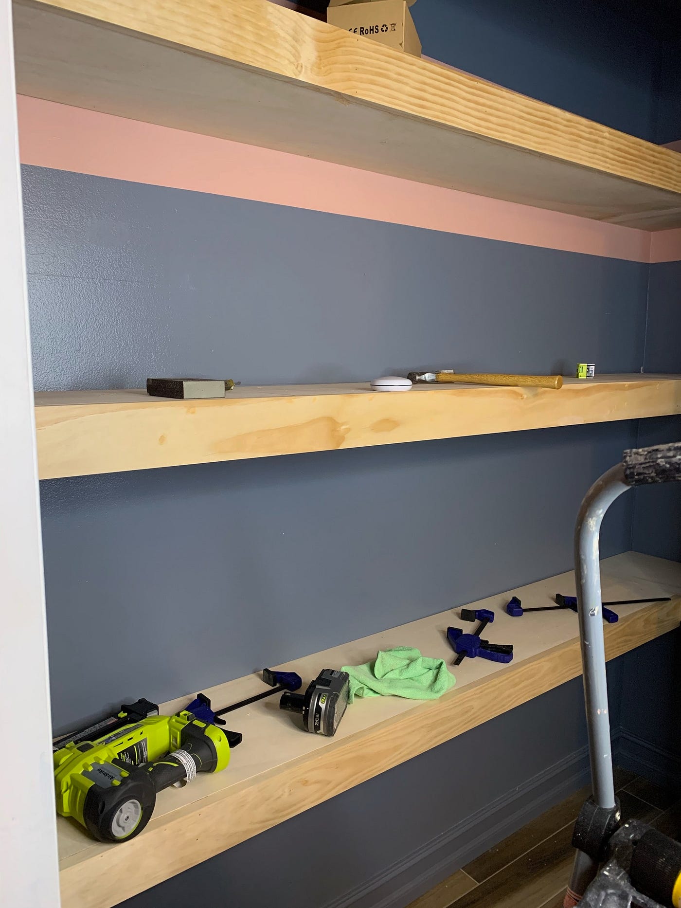Inside the closet while it was being build. There are two shelves. The top shelf has a hammer on it and some misc objects. The lower shelf has clamps, a Ryobi brad nailer and a battery. The wall is a blue/gray with the top part of the wall painted pink. The floor is a wood look tile.