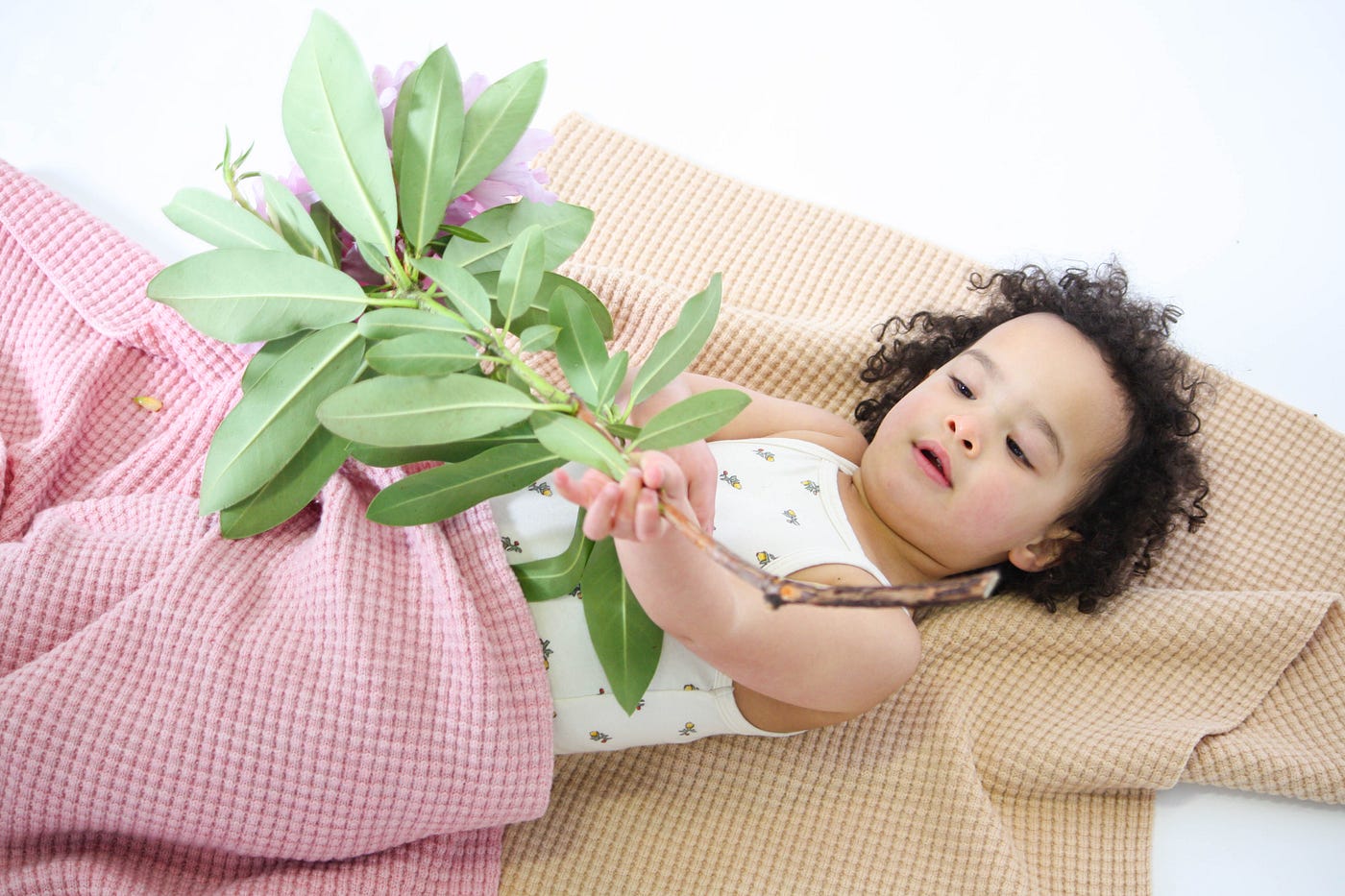 A toddler with curly brown hair holds a leafy branch while reclining with two cozy blankets.