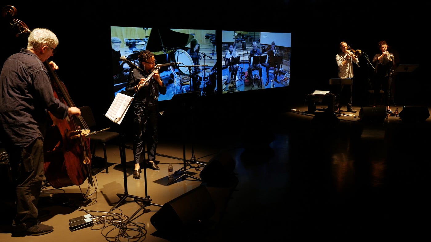 A photo of a netorked concert, with musicians on a stage joined by others on screens.