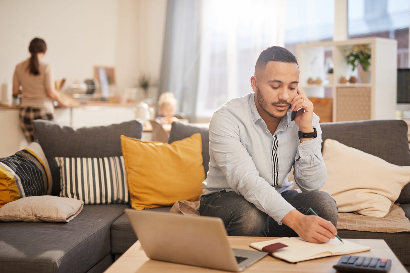 Man sitting on a couch talking on a cell phone whilst taking hand-written notes in a journal.