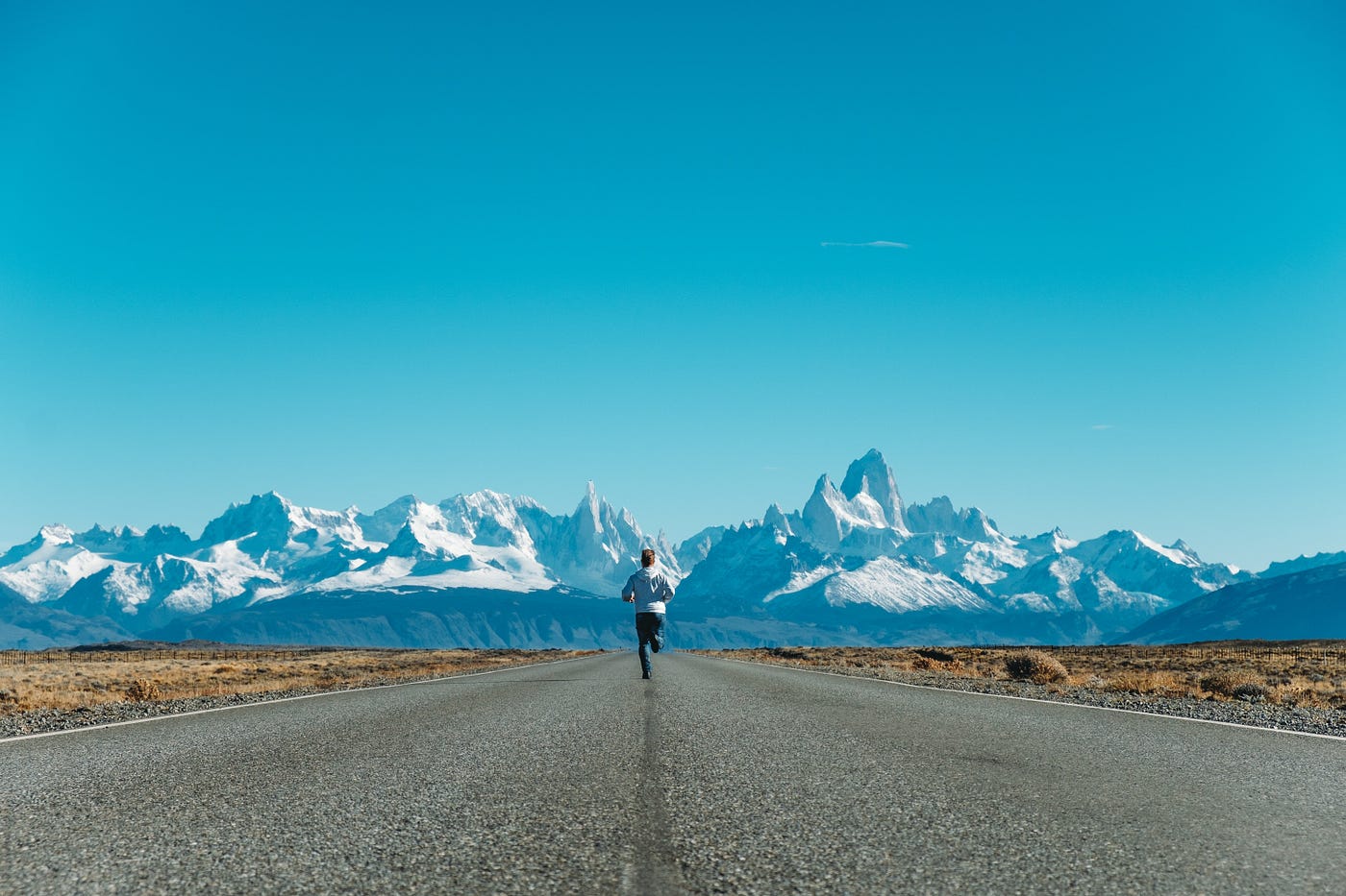 A man running along a wide, straight road toward distant, snow capped and jagged mountains