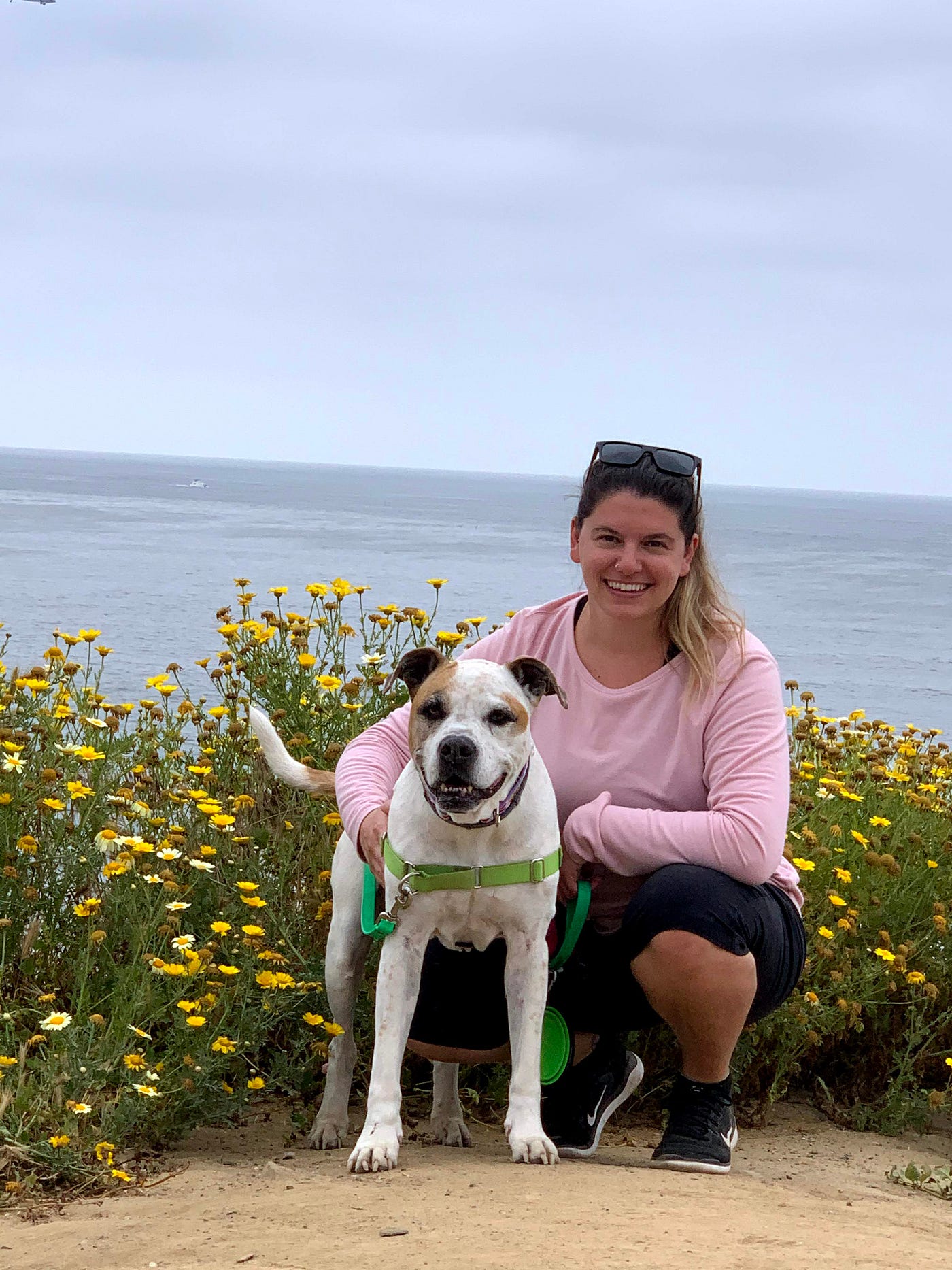 Sandra with her dog that looks like a pitbull mix in front of an ocean and yellow flowers