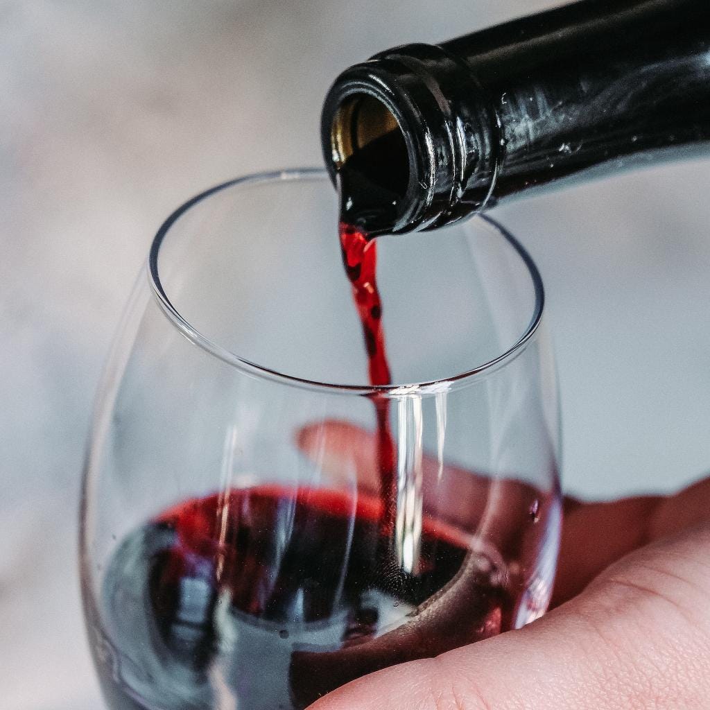 A relatively close image of red wine being poured from a wine bottle into a wine glass being held by a hand.