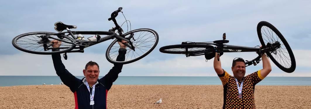 Two men standing on a beach holding their bikes above their heads, with the sea and a sea gull in the background