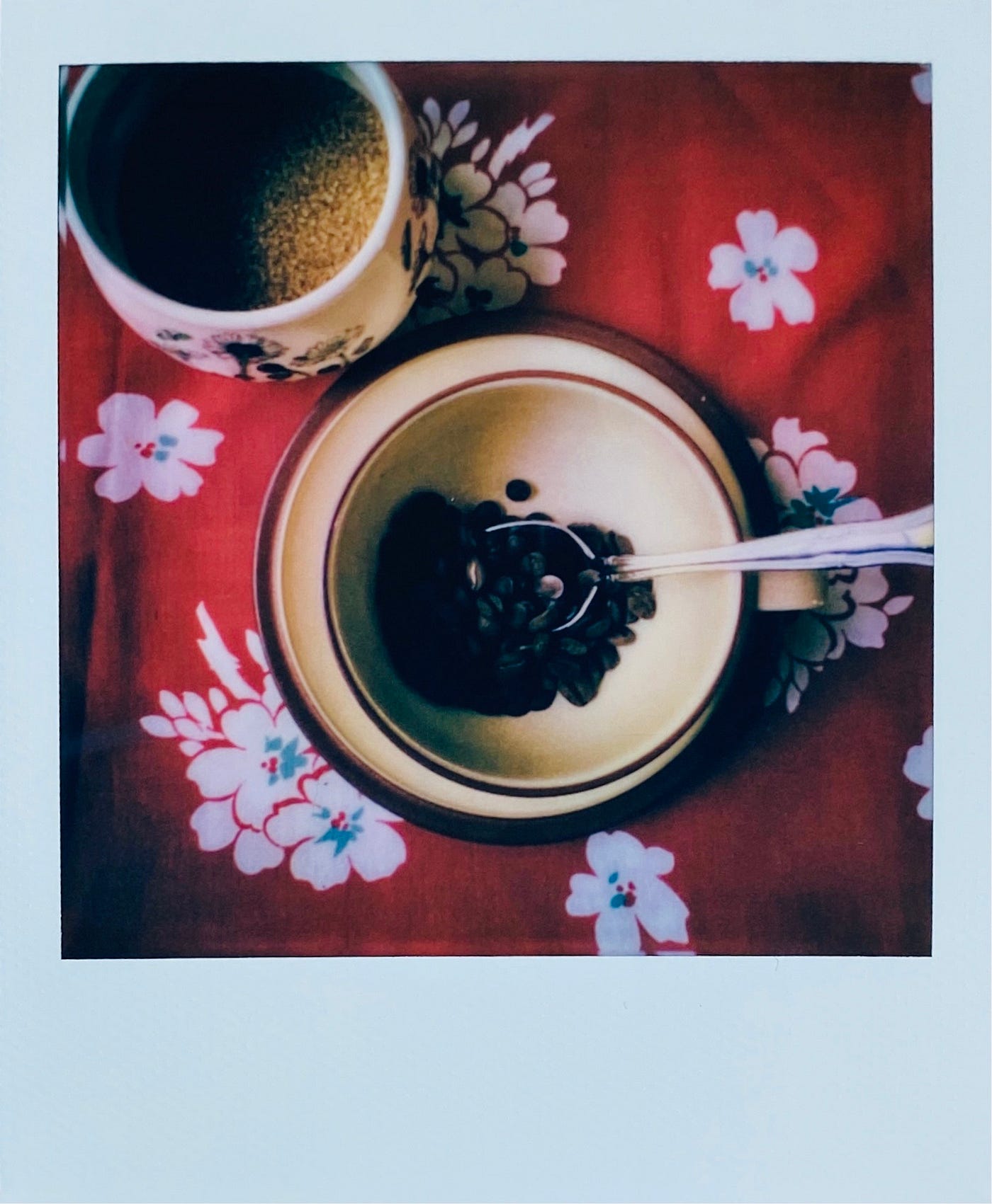 A coffee cup with coffee beans inside, resting in a spoon, and a sugar bowl with raw sugar inside, on red & white background