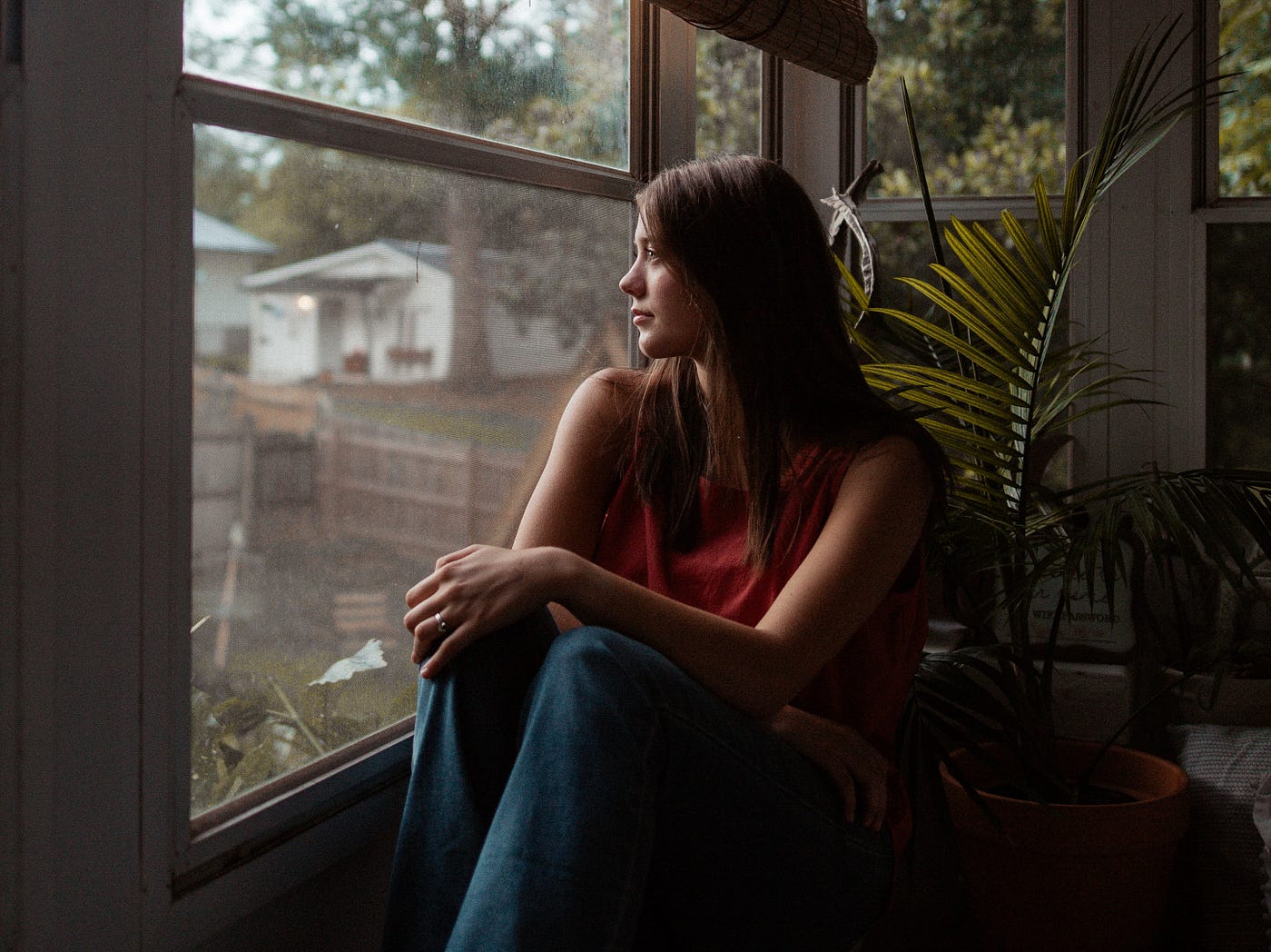 Woman sitting and looking outside window with plants behind her. She’s wearing jeans and a red shirt.