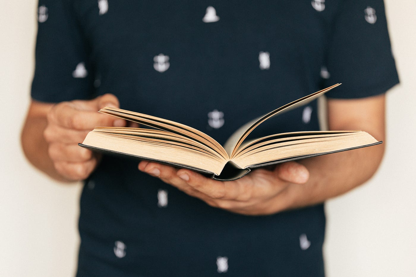 A man in a navy blue shirt dotted with white holds an open, cream-colored book in both hands.