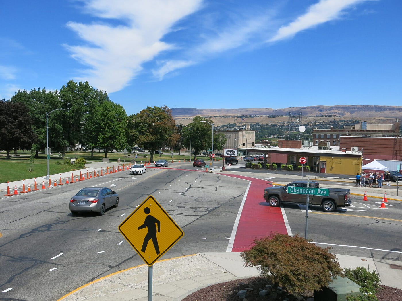 una vista elevada de una intersección de cuatro vías con autos circulando. Una señal de paso de peatones en primer plano y los pasos de peatones están pintados.