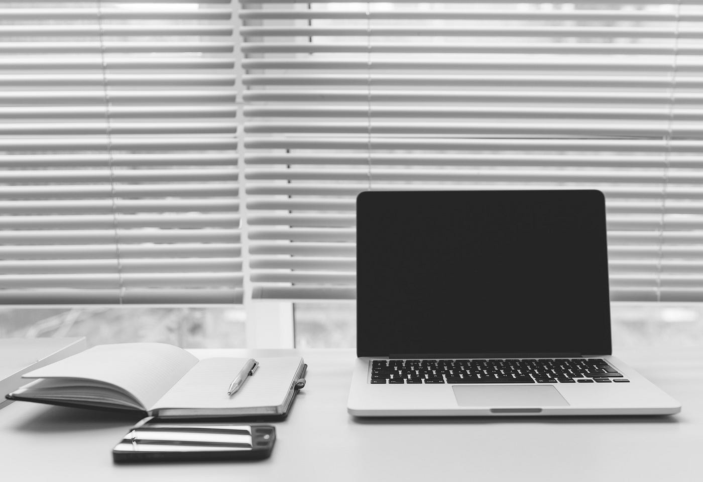 Desk with empty, open notebook with a pencil on it to the left, a smartphone head down and an open laptop to the right.