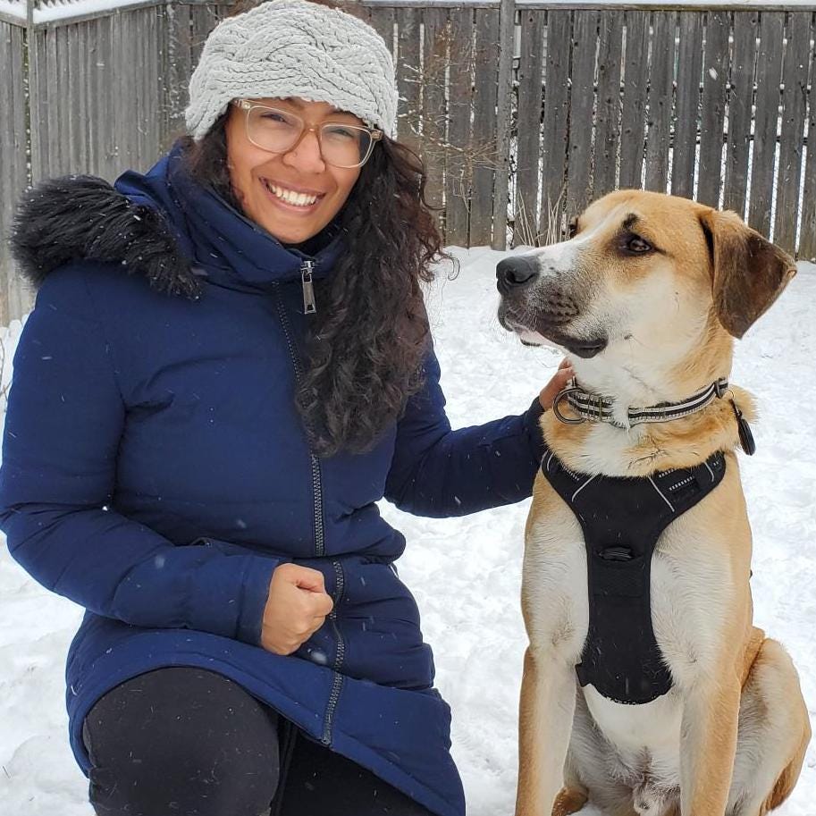 Catherine smiles and kneels next to Mickey (a beige and white medium-sized dog) in the snow