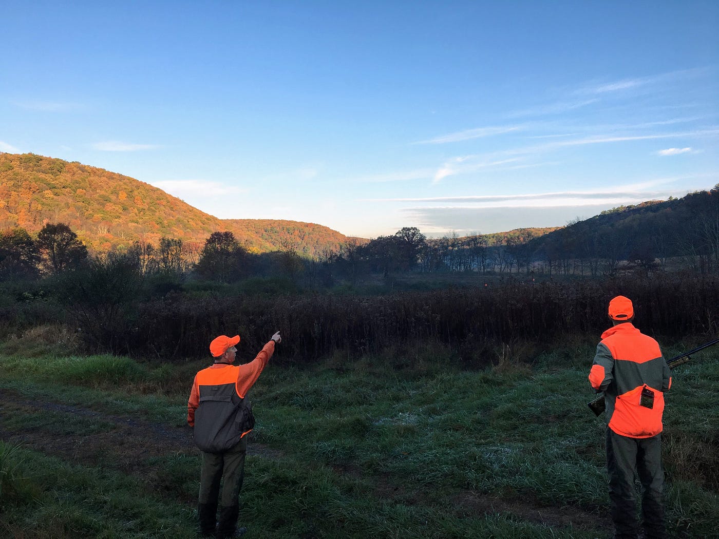 Two men, wearing fluorescent orange hunting clothes, stand on a grass road. One points to a field as the sun rises over the valley they’re in.