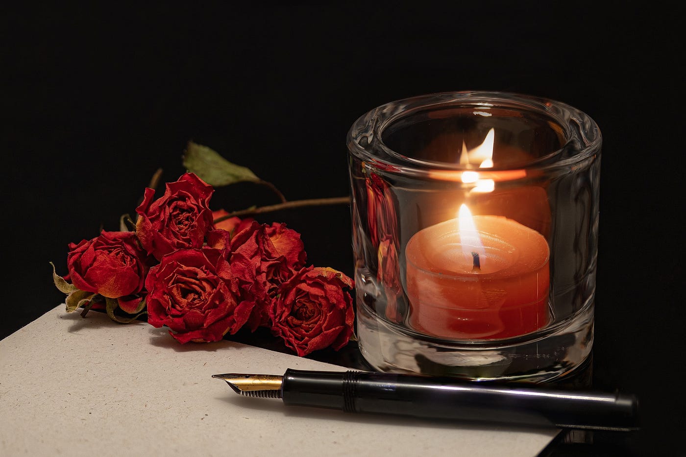A burning candle within a glass holder, next to a fountain pen and dried roses.