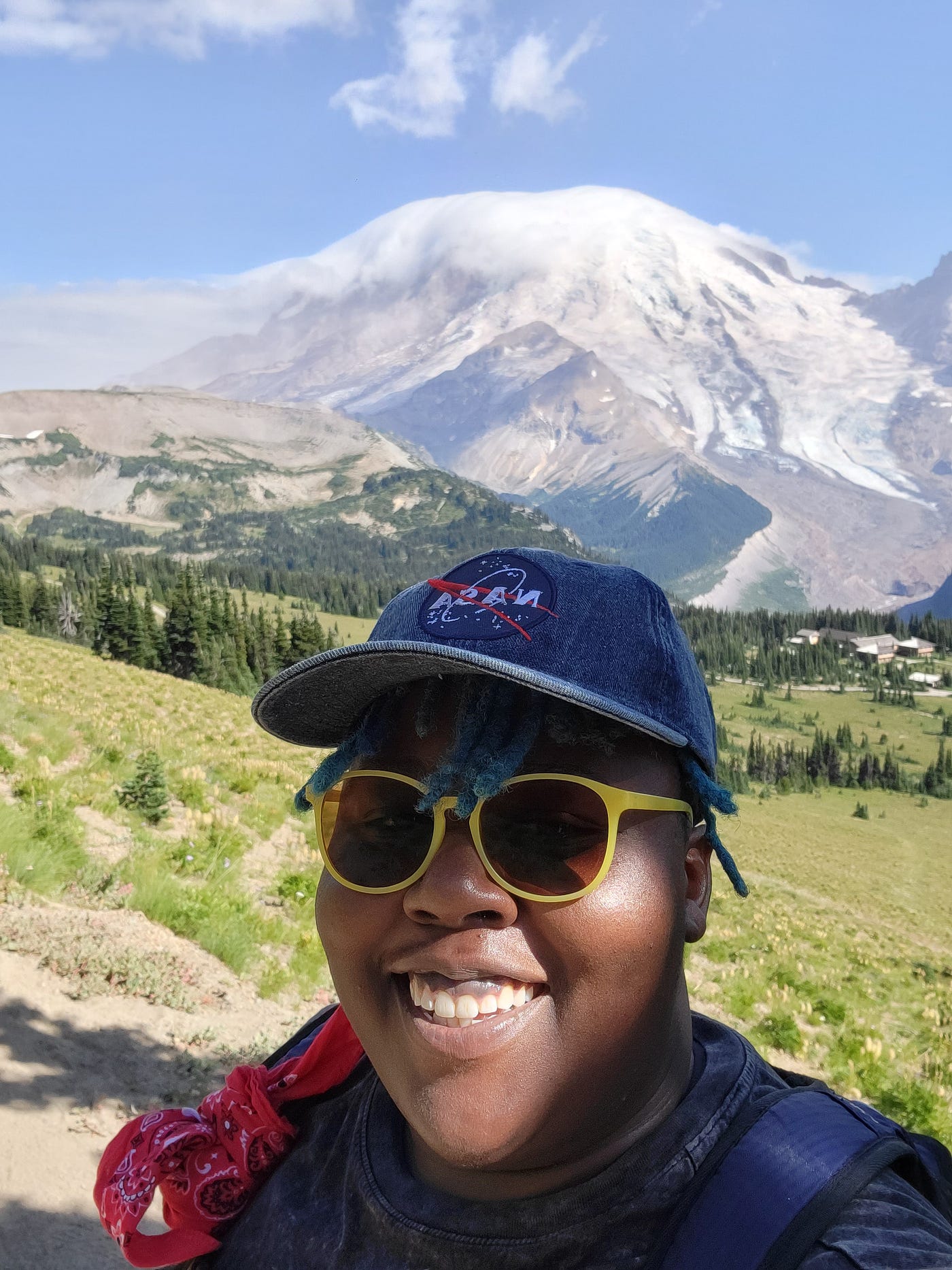 Author on top of a mountain with green hills rolling behind them leading toward a mountain top crested with clouds on a sunny day. Aigner Loren Wilson is smiling wearing yellow sunglasses and a jean NASA hat. Author’s life. Life of a publishing speculative fiction author. Speculative Fiction author. Aigner Loren Wilson
