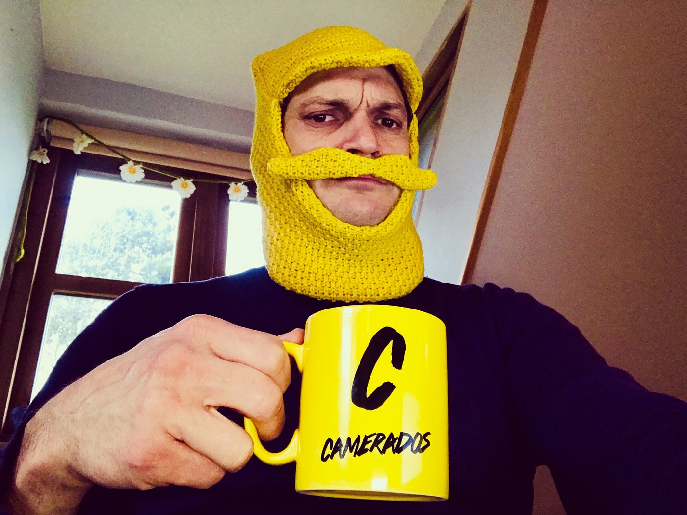 A man wearing a yellow hat that has a moustache holds up a yellow mug that has ‘Camerados’ on it.