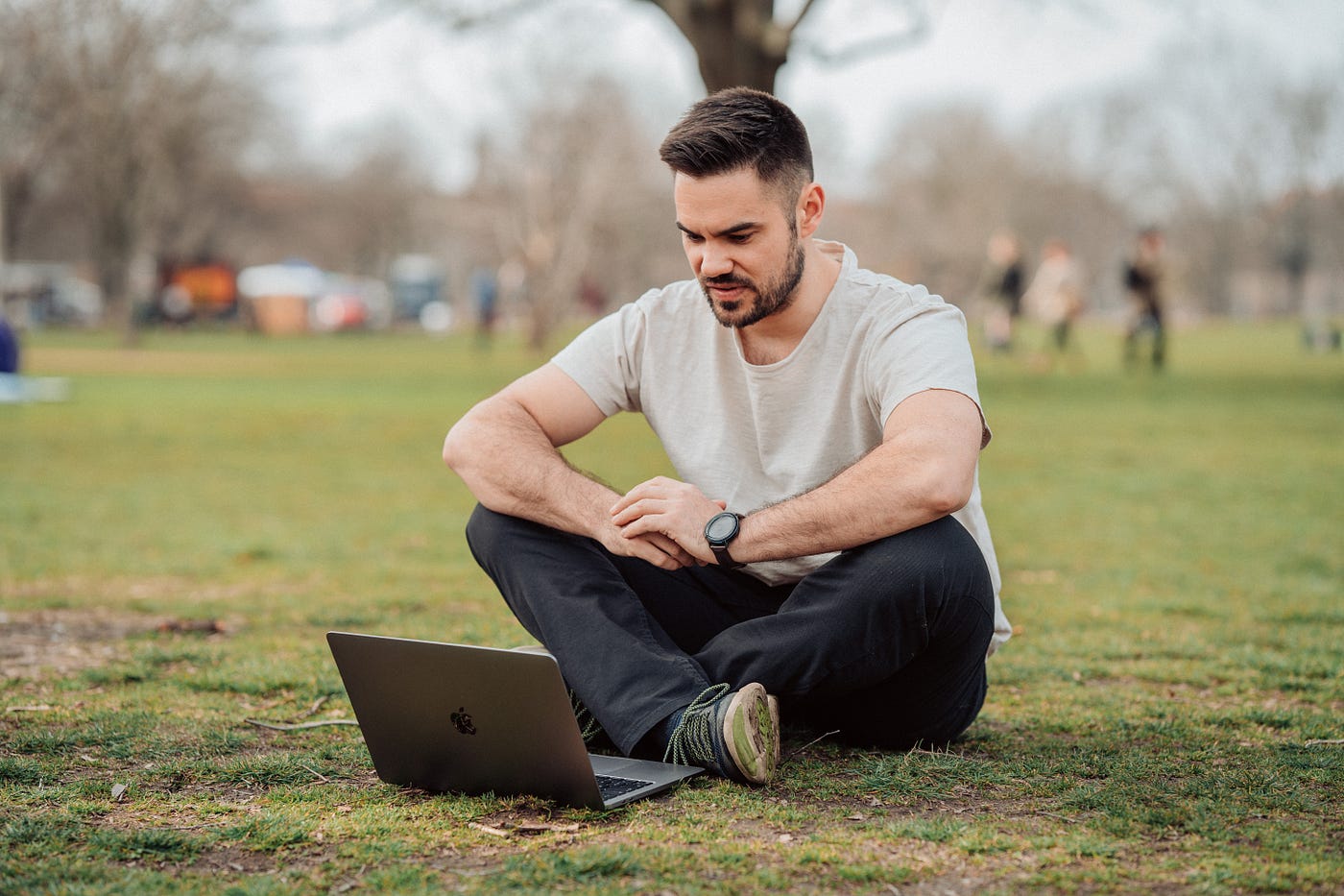 A man sitting behind his MacBook on the ground on a park