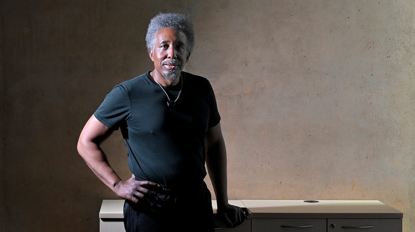 Frank B. Wilderson III stands with his right hand on his hip and his left hand resting on a table
