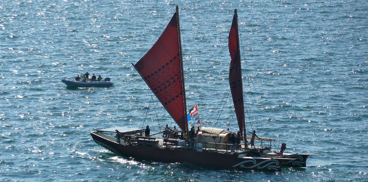 The waka Haunui floating on a rippled ocean with a small inflatable boat in the background. The waka is a replication of the style of canoe that early Māori may have used to voyage to Aotearoa New Zealand.