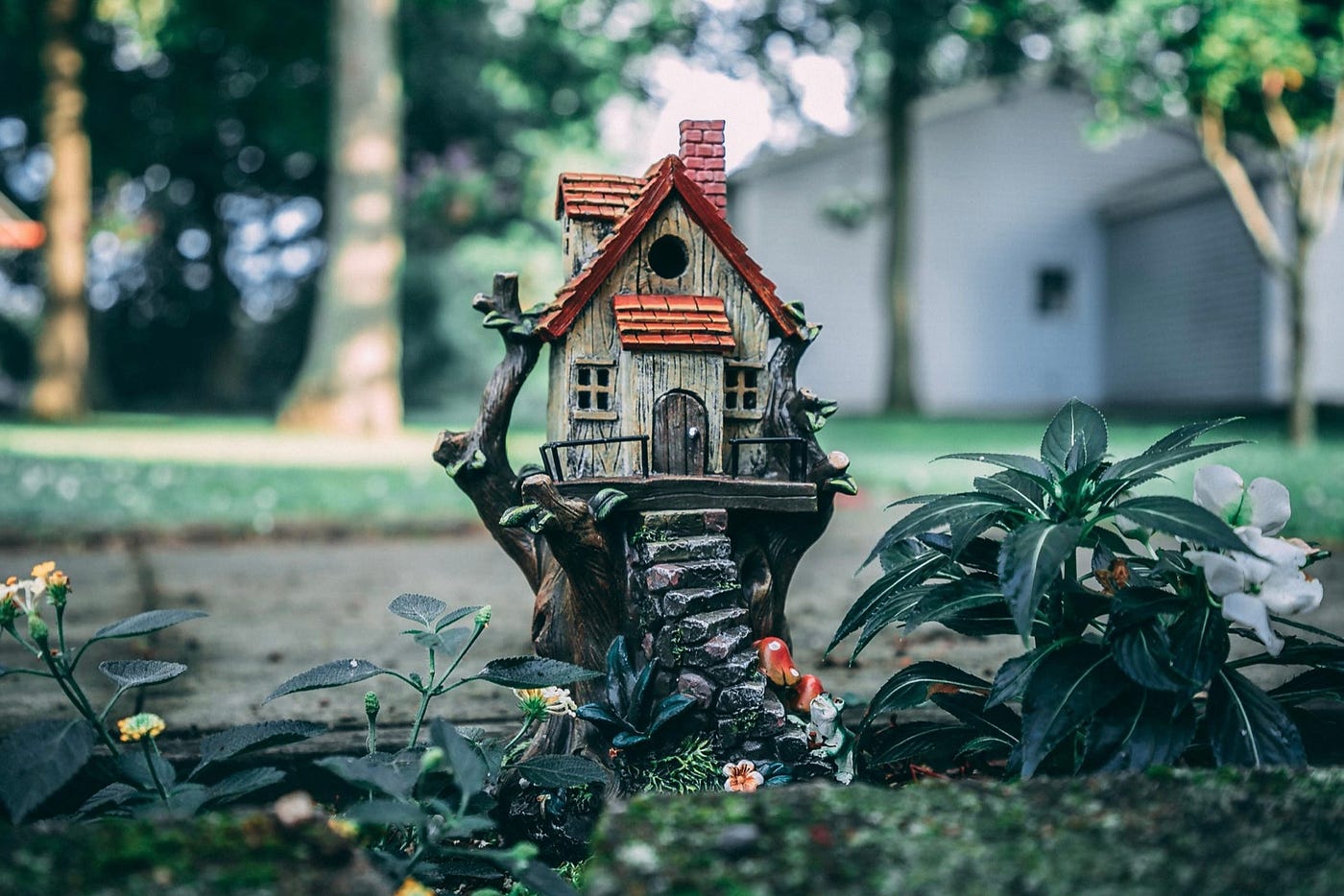 A garden figurine of a small cottage with winding concrete stairs is in the center of the photo. The room is tiled in desert orange and there is a brick chimney to the back right. The siding is unsealed wood planks and the house sits on the trunk of a tree. The figurine sits by an white impatiens flower and other small flowers. Big trees and a household are seen in the background.