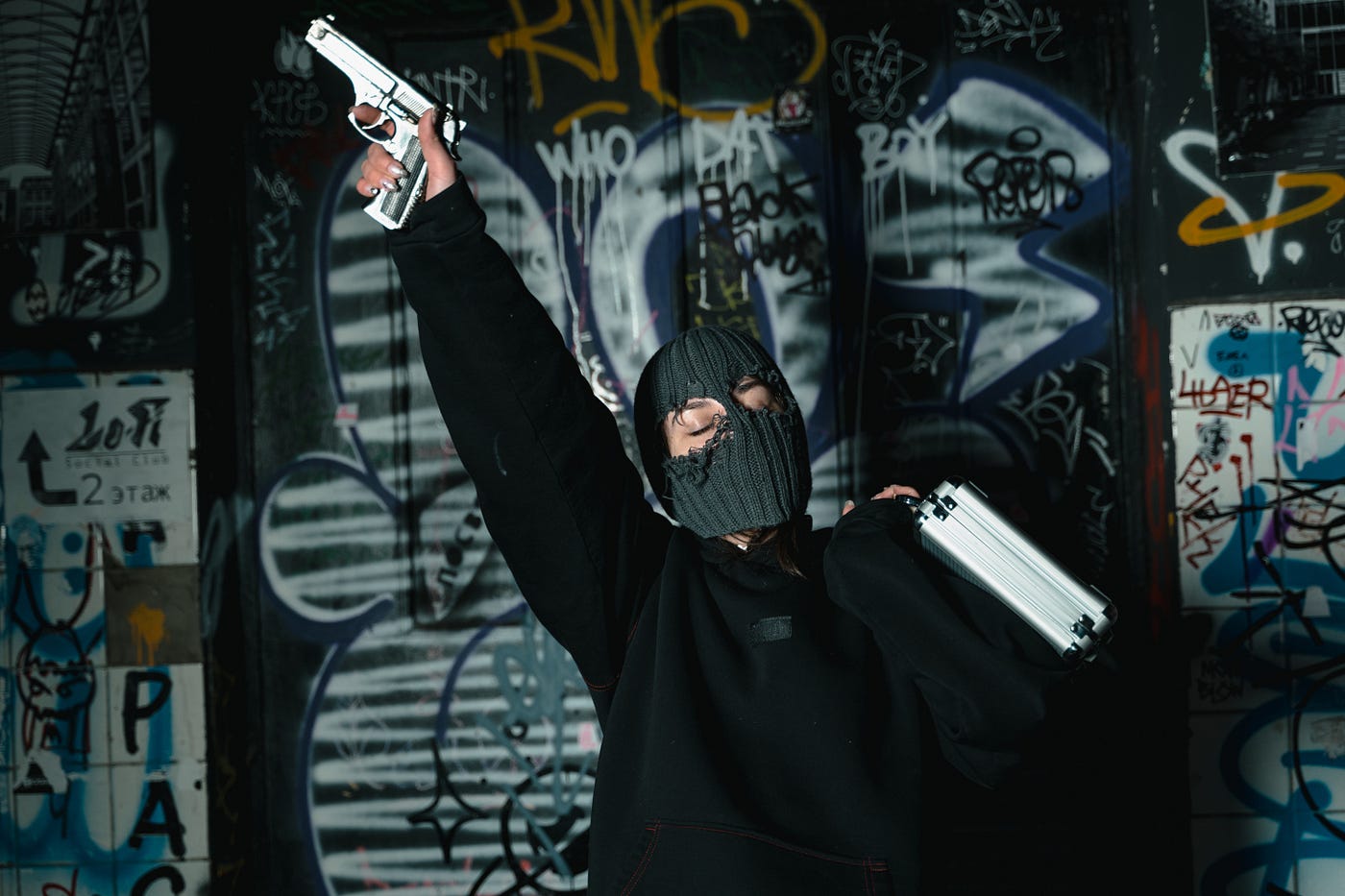 A woman wearing a balaclava and pointing a gun up, with a suitcase in her hand. There is graffiti in the background.