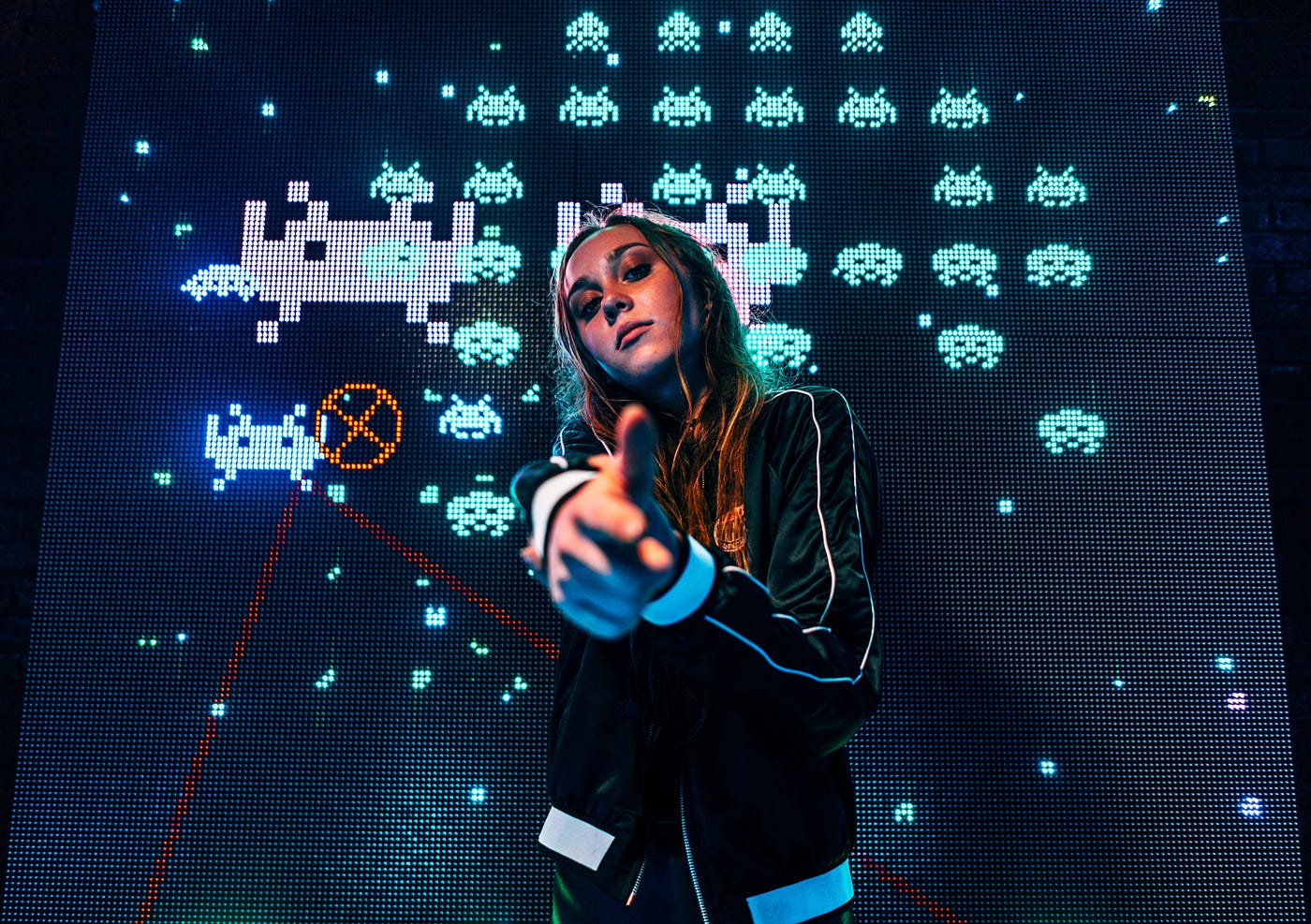 Girl making finger-guns at the camera, standing in front of a screen showing gameplay from ‘Space Invaders’