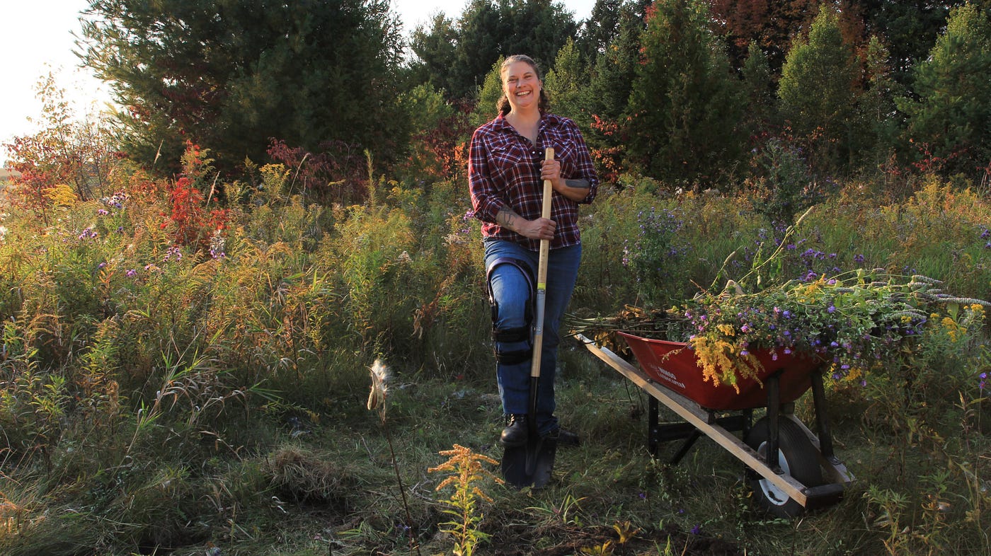 A white disabled Jewish genderqueer femme in a wildflower field with a shovel &smiles at the camera,next to a red wheelbarrow
