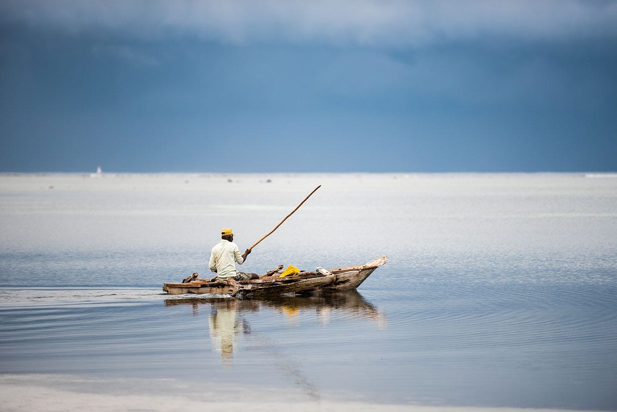 A lone fisherman on the water in a very small, wooden boat. He is holding a fishing line. There are dark, storm-like clouds in the distance.