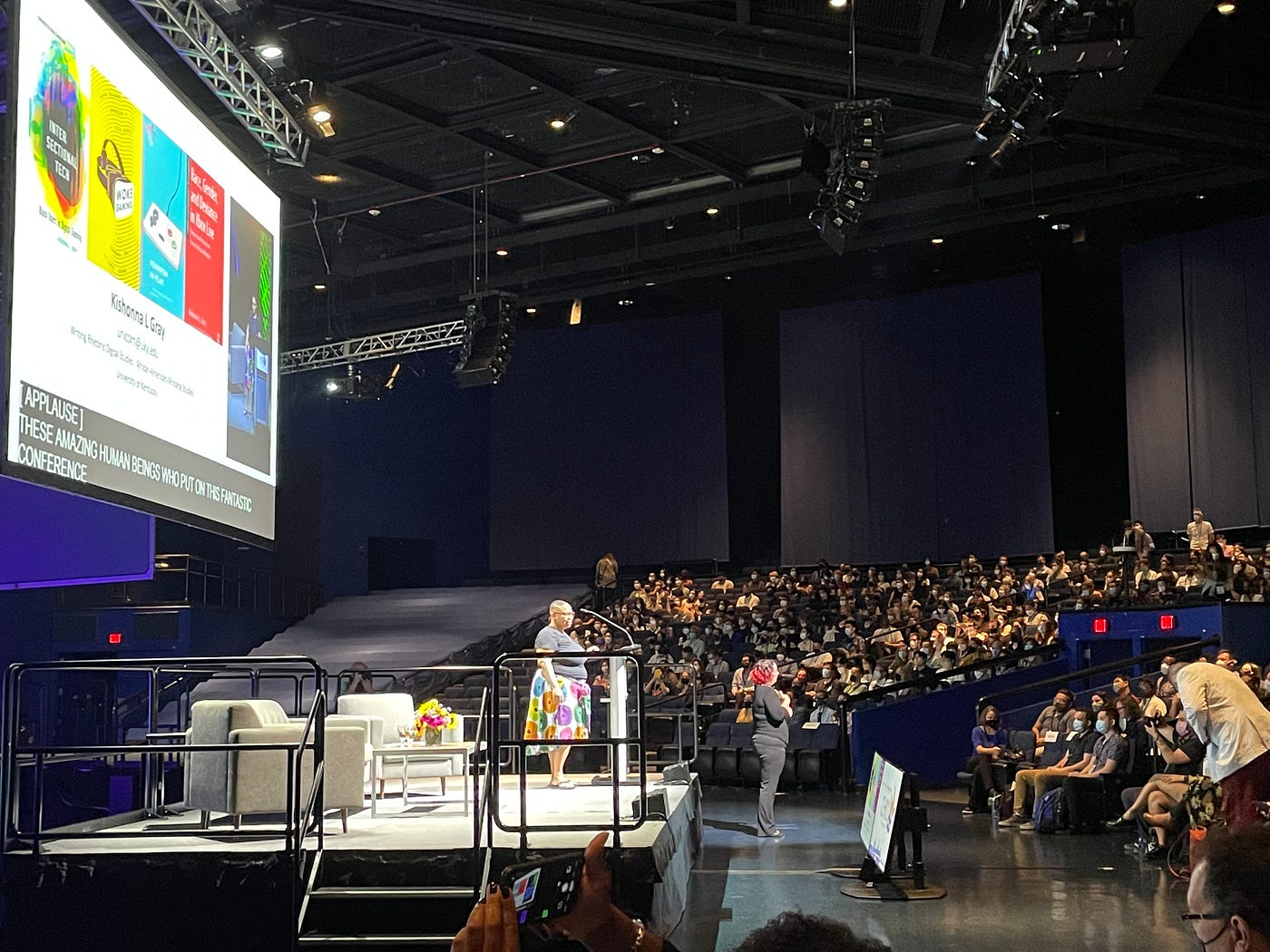 Kishonna in a black top and colorful skirt on stage with couches and a presentation slide showing a medley of game images.