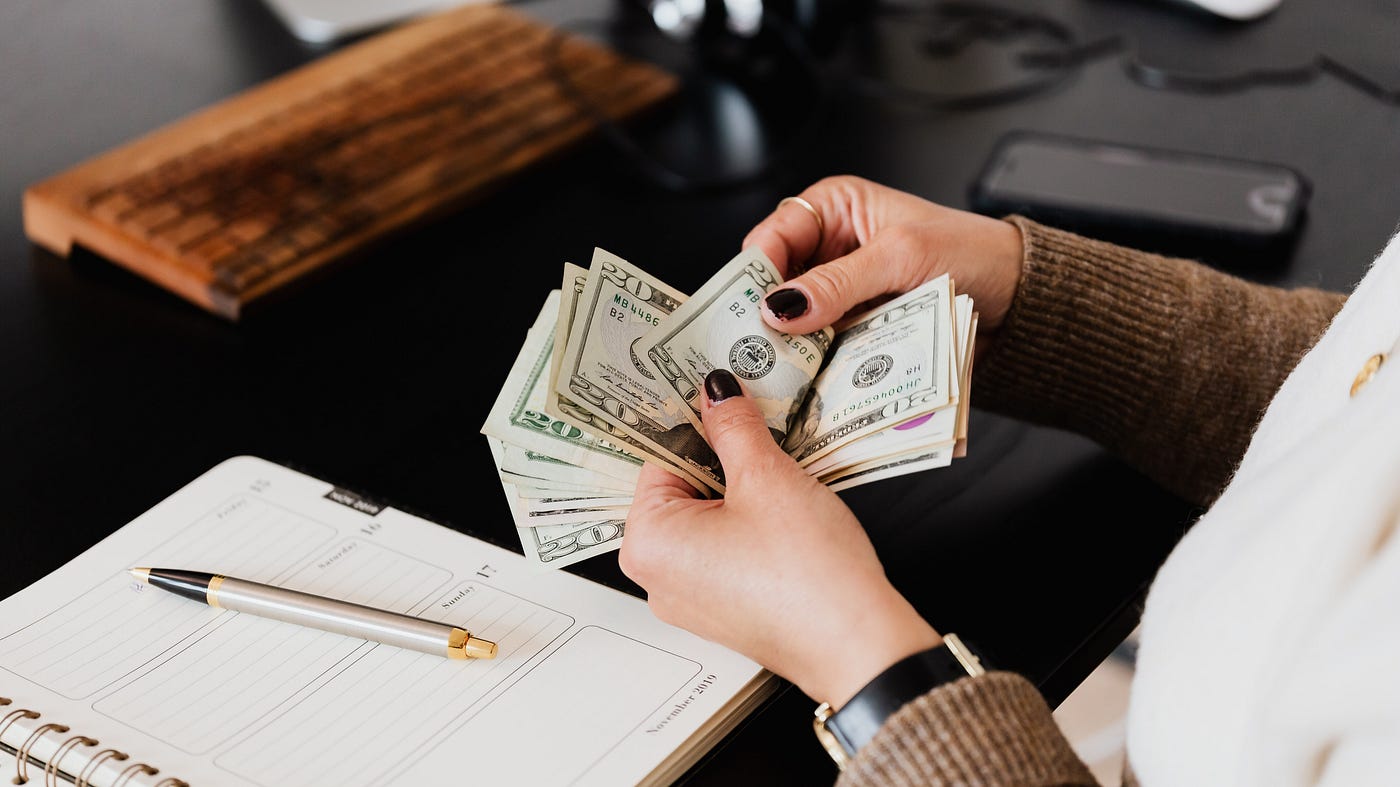 Womans hands holding a stack of $20 bills. A pen rests on top of a notebook on the desk in front of here.
