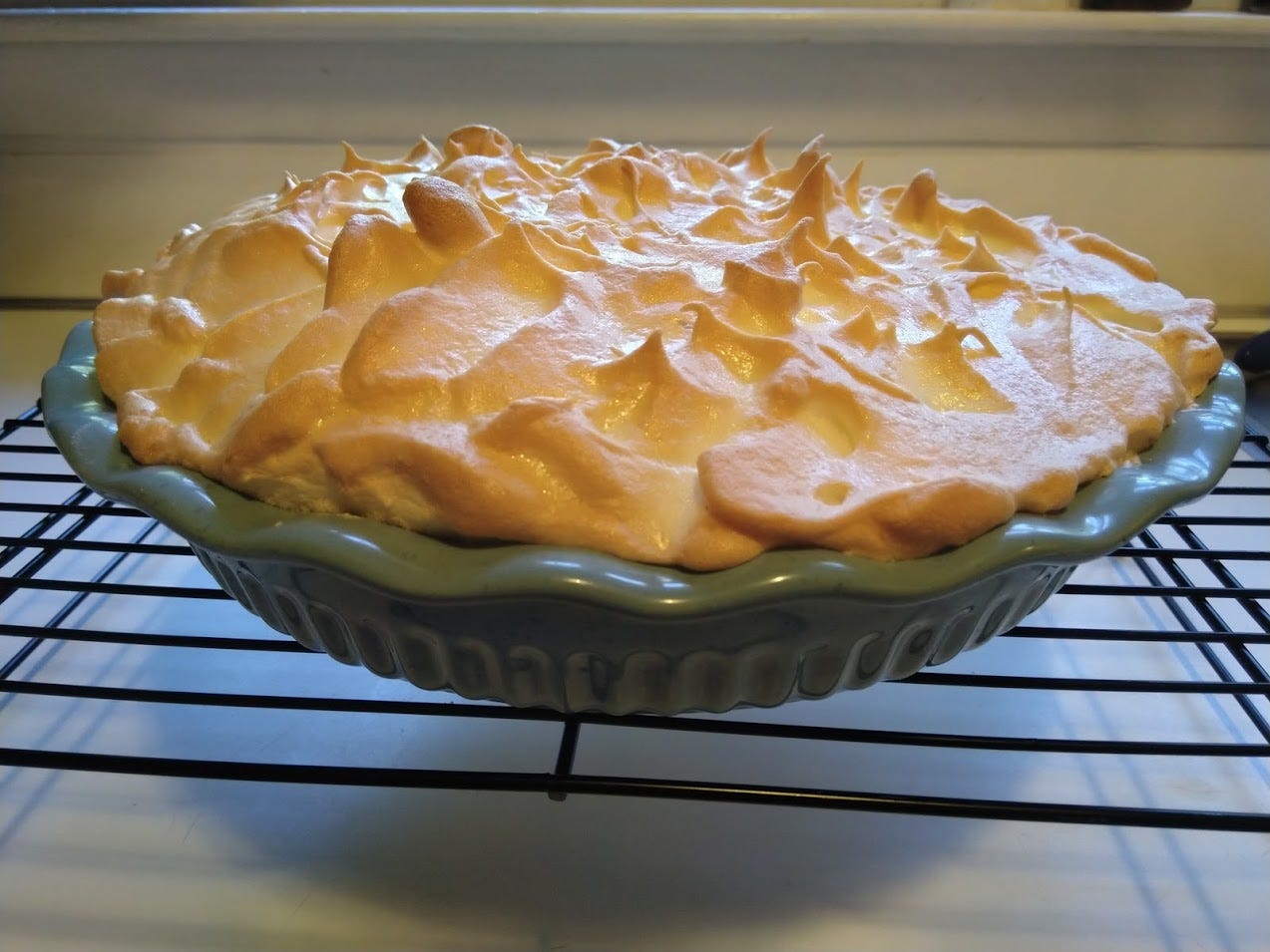 A golden brown merangue pie in a teal ceramic pie dish, cooling on a rack