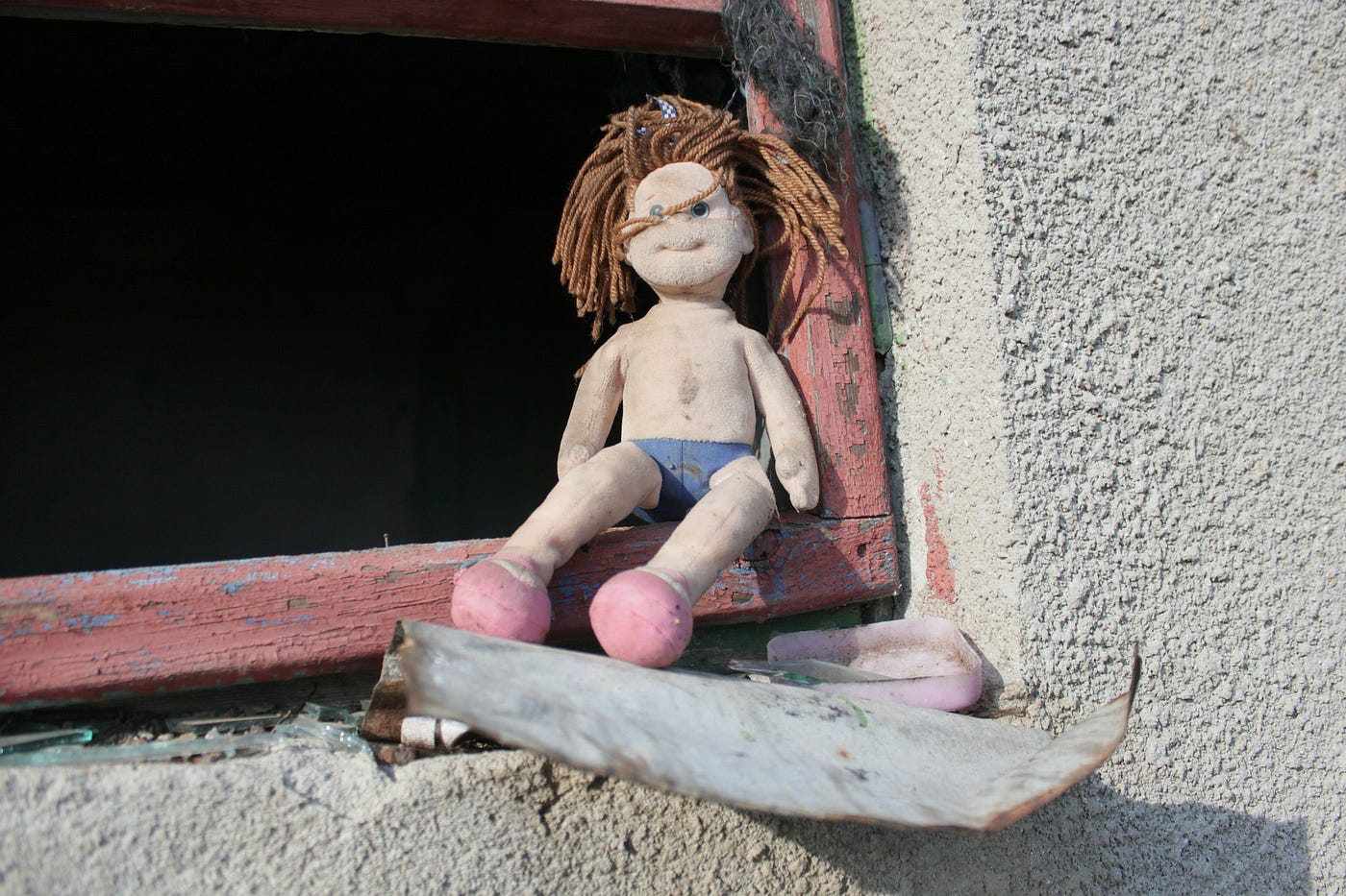 Kill, Let Kill, Or Be Killed. A small ragdoll sits abandoned, unclothed, and dirty in the corner of a glassless window with broken concrete around the pane.